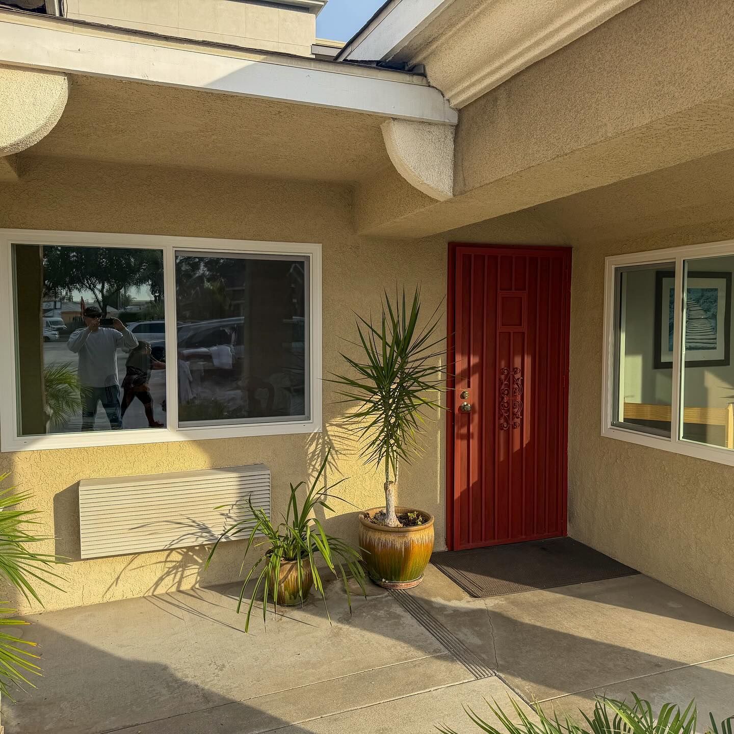 Front entrance of a beige house with a red door, large window, and potted plants in sunlight