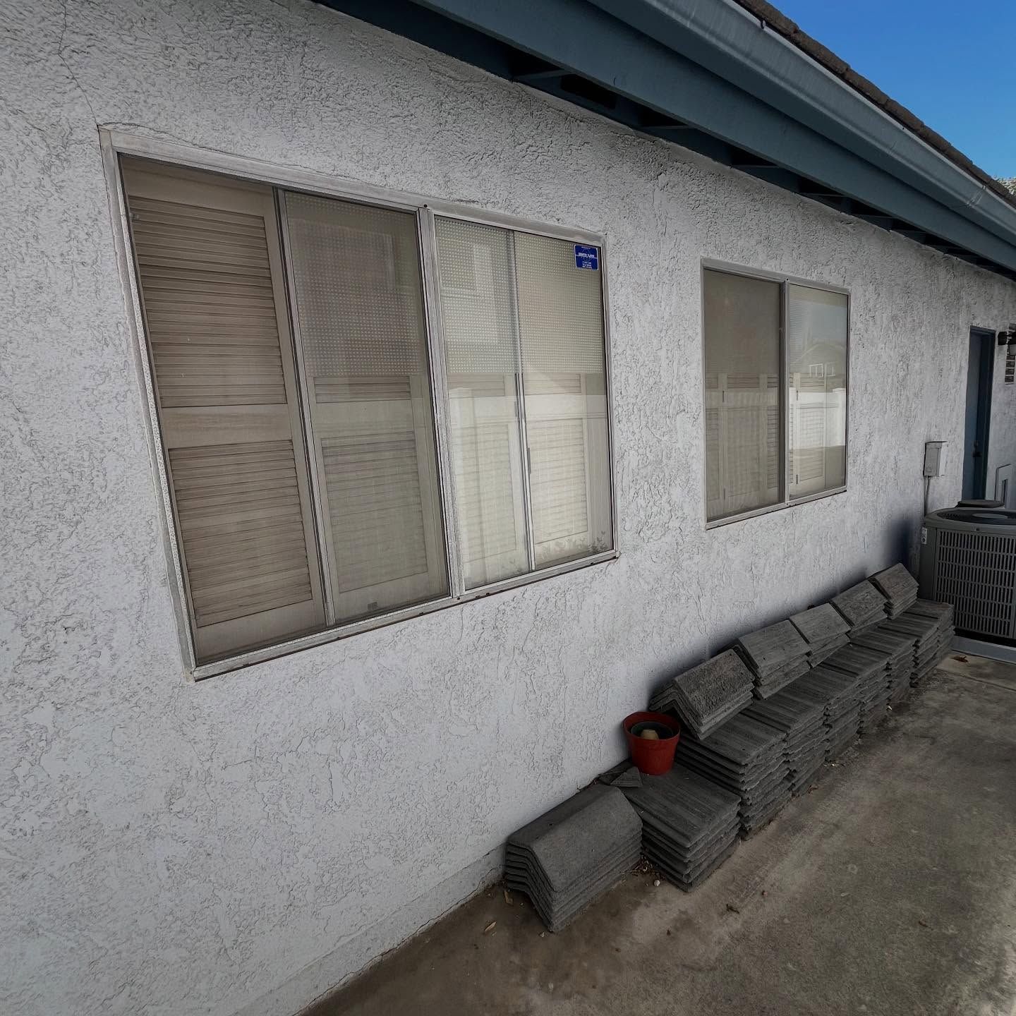 Exterior stucco wall with windows, stacked concrete blocks, and a red bucket on a patio under an eave