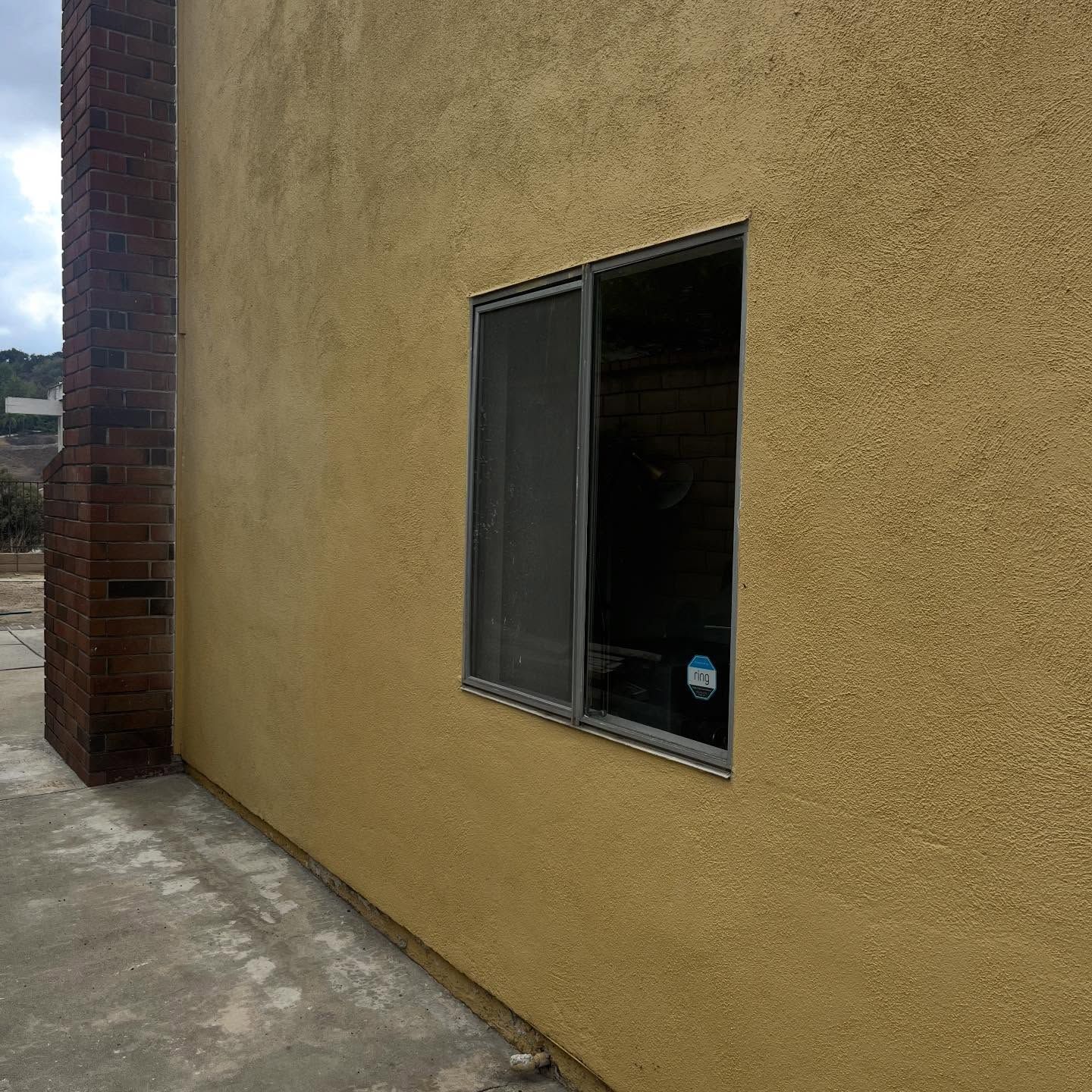 Yellow stucco wall with a small open window beside a brick corner on a concrete walkway