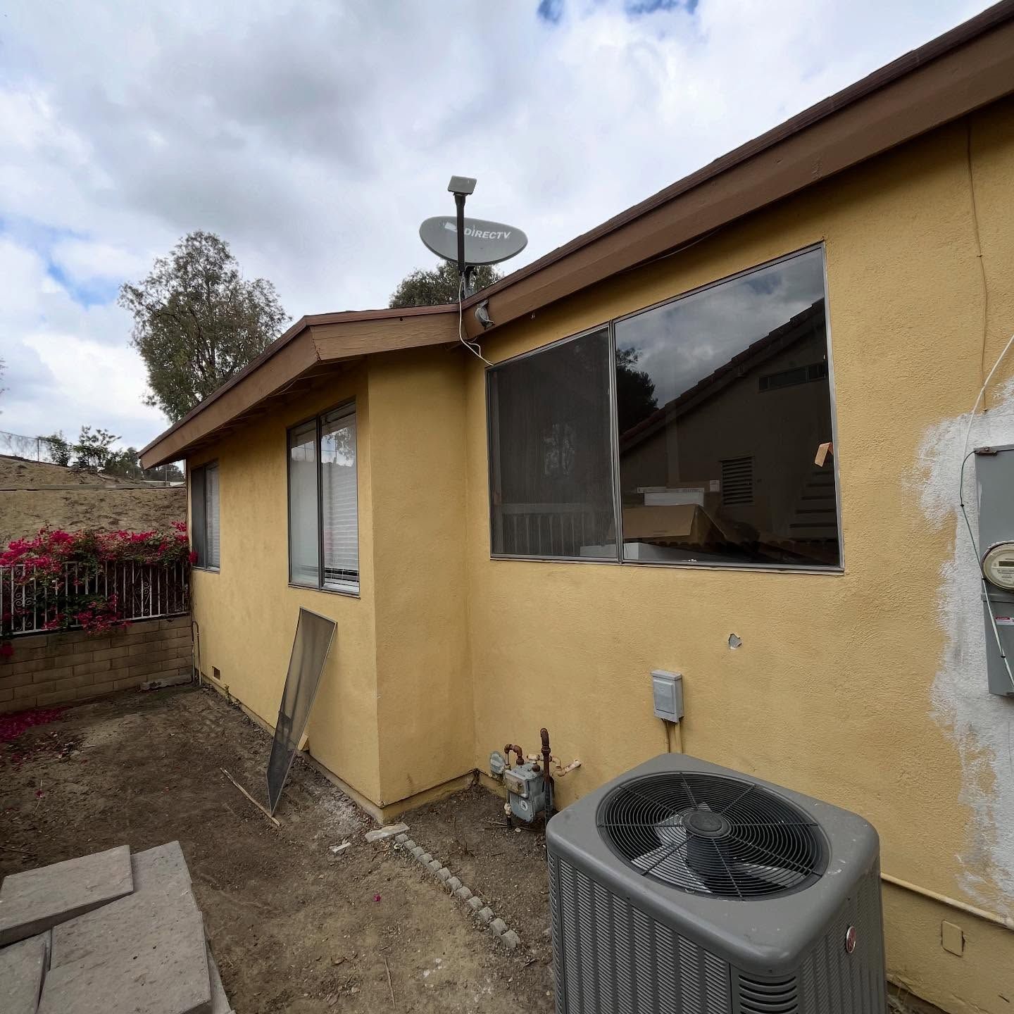 Backyard side of a beige house with windows, dirt ground, and an outdoor air conditioner unit