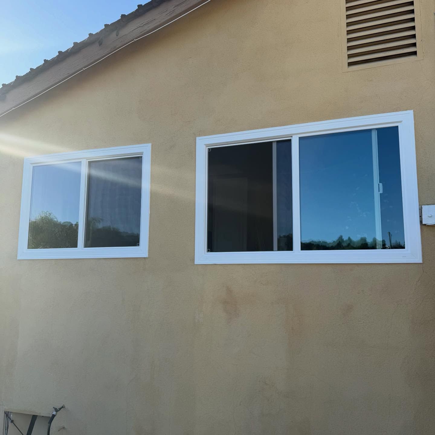 Beige house exterior with two white-framed windows and a roof vent in sunlight