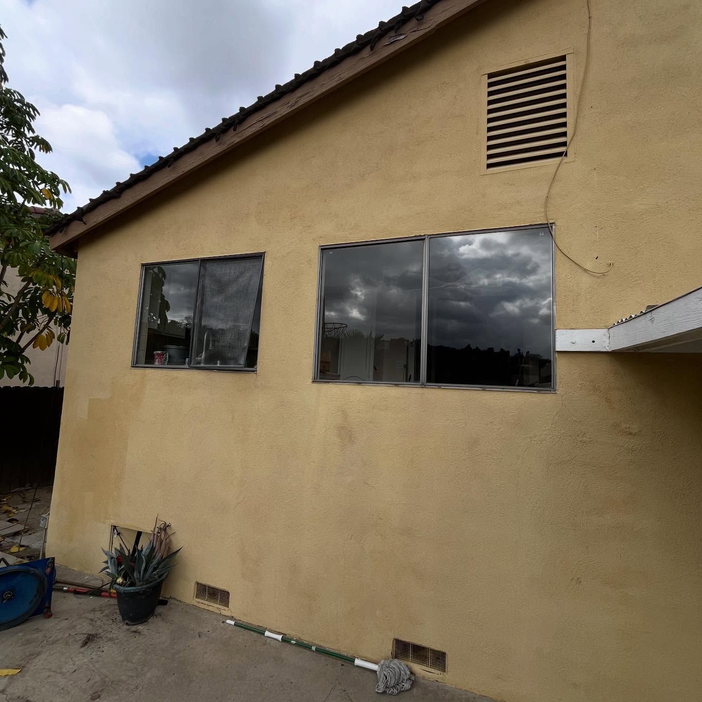 Yellow stucco house exterior with two windows and a small vent under a cloudy sky