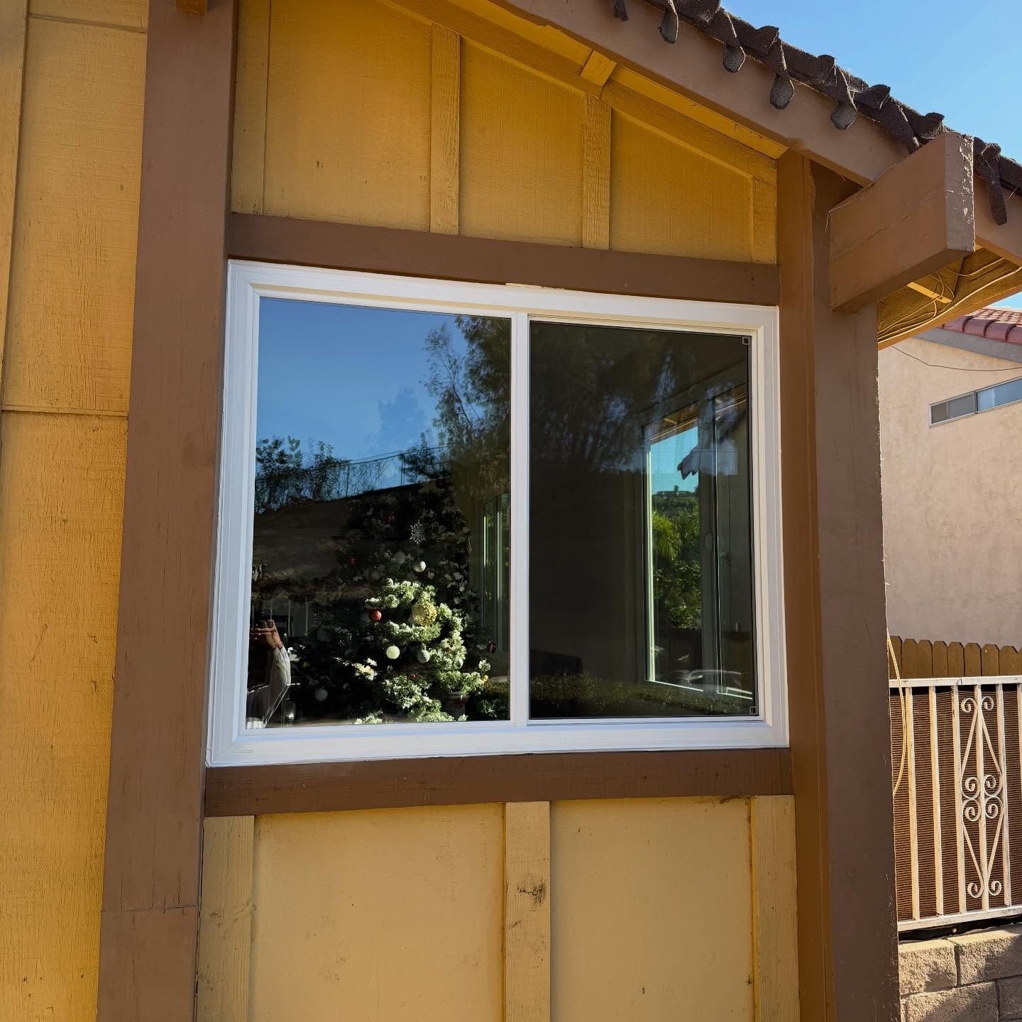Small window in a tan and brown house exterior with white trim and a reflected tree.