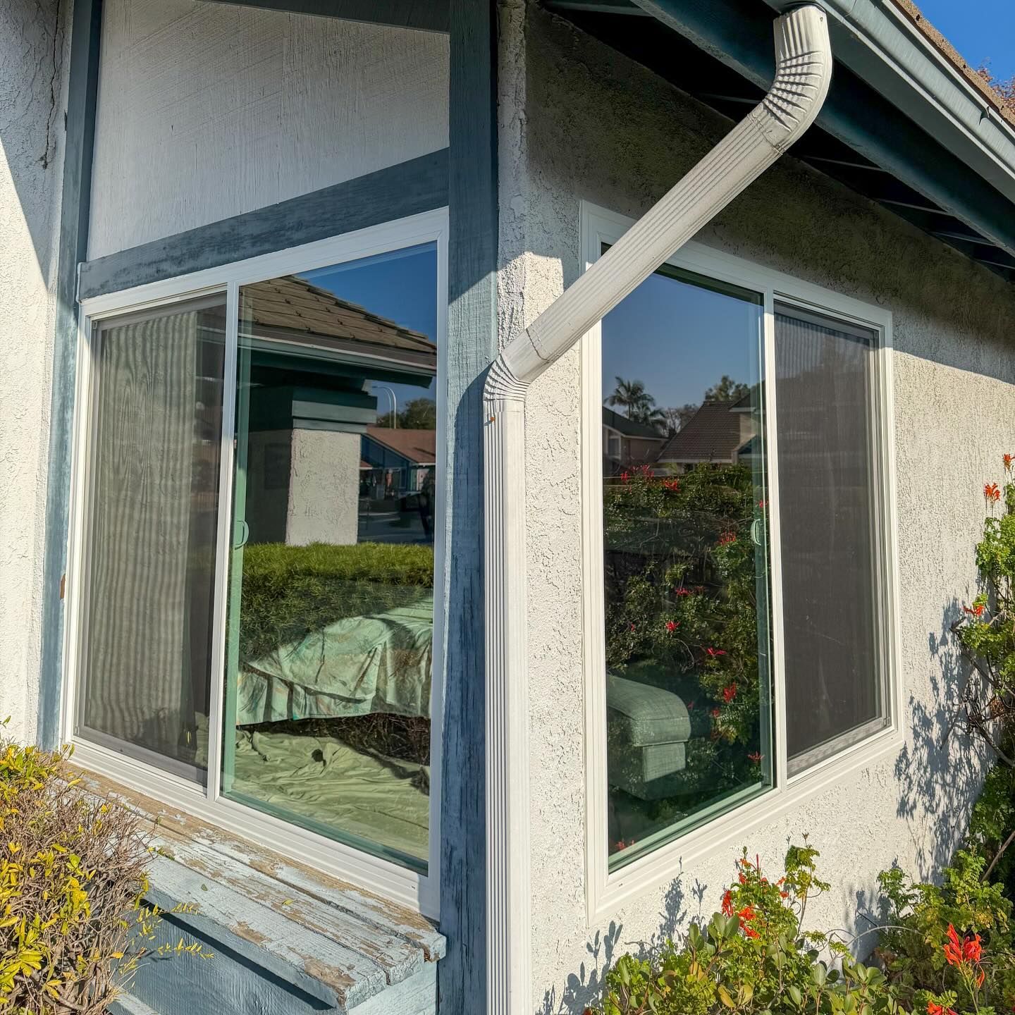 Corner of a house with large windows, white stucco walls, and a downspout, beside a garden with flowers.