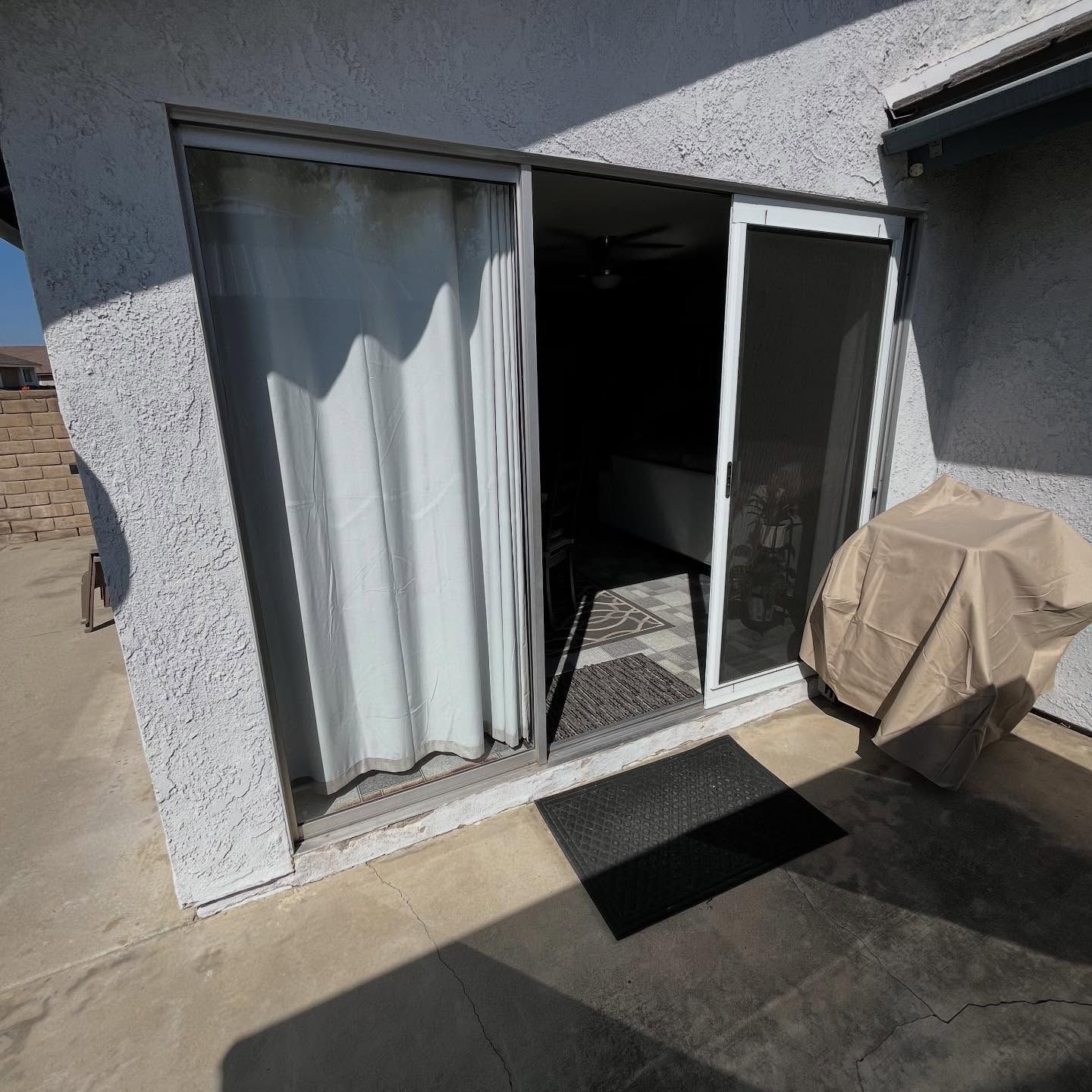 Sliding glass door to a patio, with sheer curtains inside and a covered grill beside it.