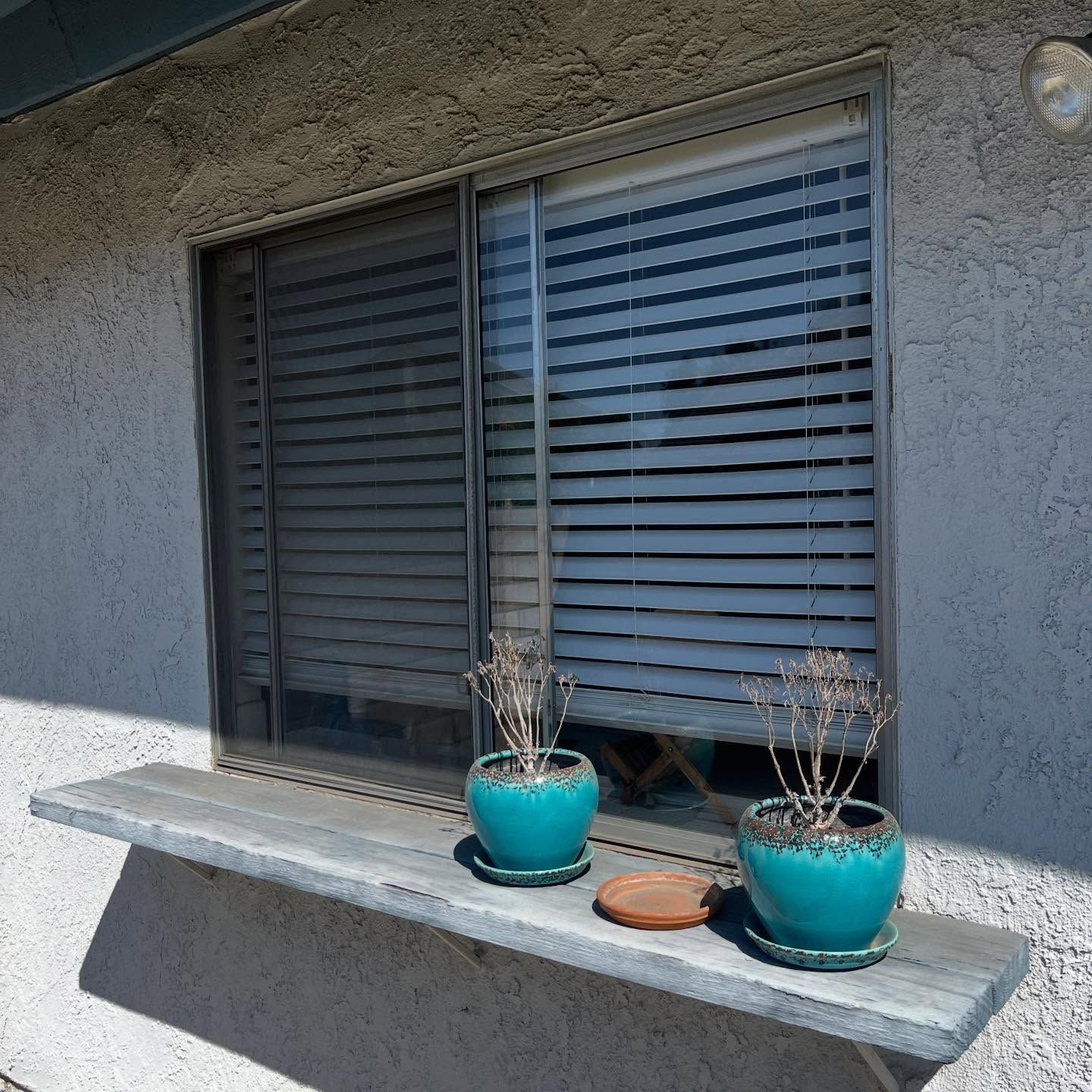Two turquoise potted plants on a concrete window ledge beneath closed gray shutters