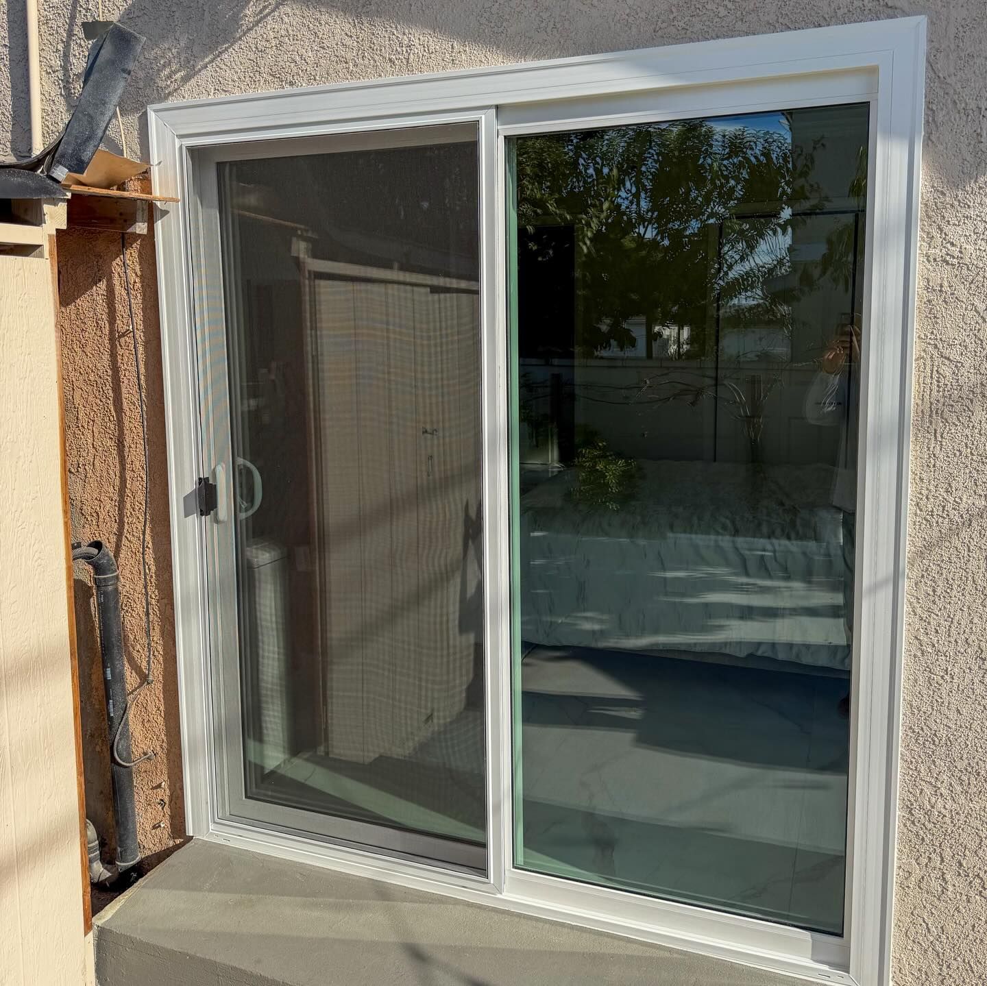 White-framed sliding glass door on a stucco exterior wall, reflecting a patio and trees.