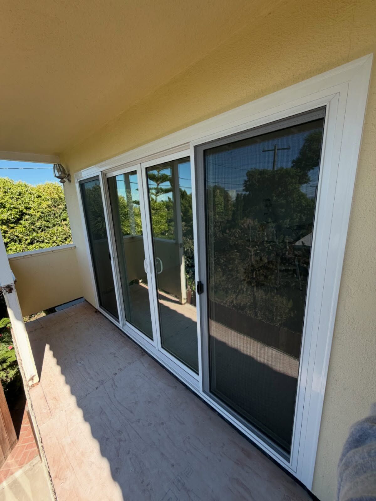 Outdoor balcony with white sliding glass doors and tiled floor, overlooking trees and a neighborhood.