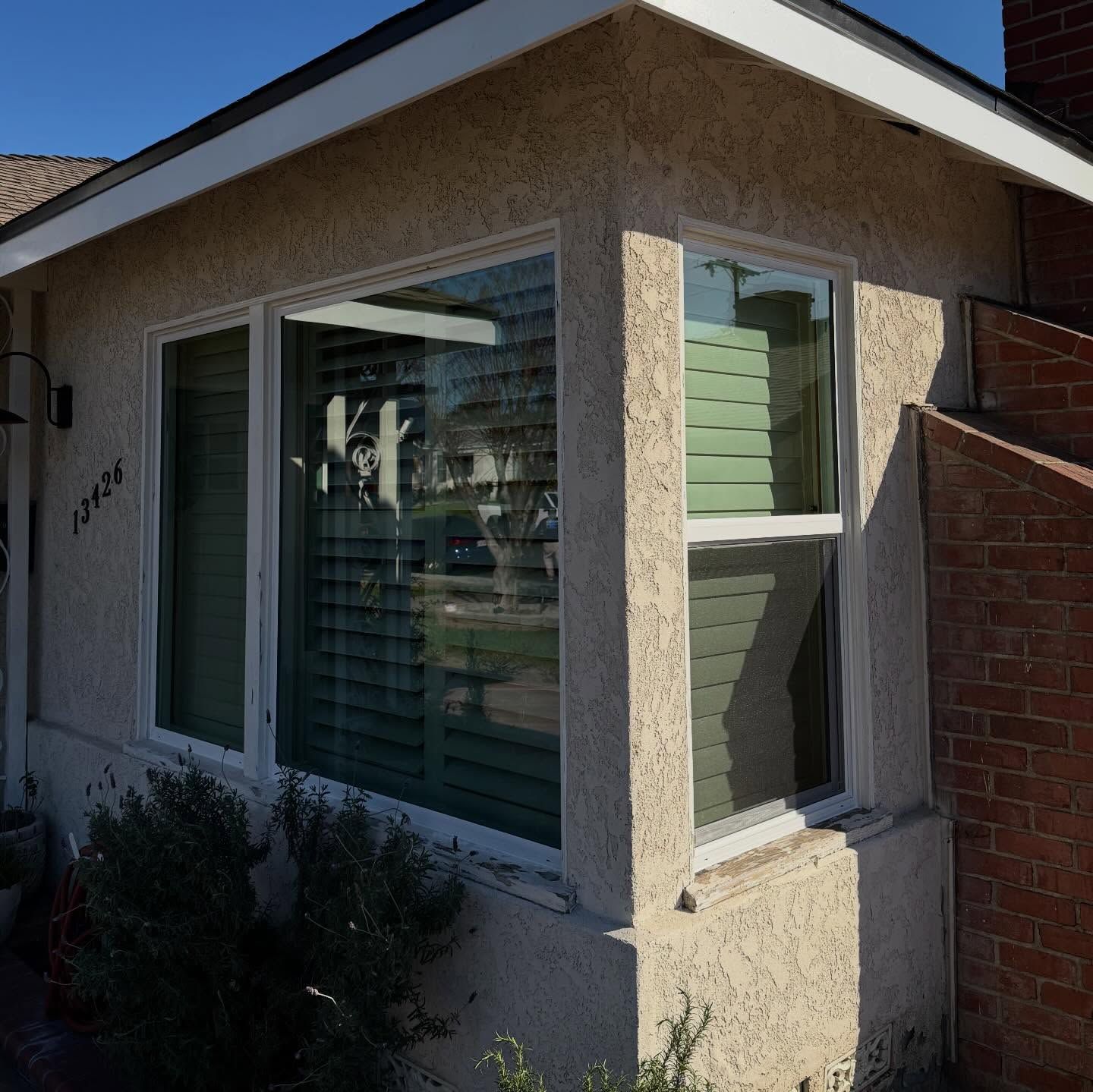 Front corner of a tan stucco house with large windows and white trim, beside a brick wall and shrubs