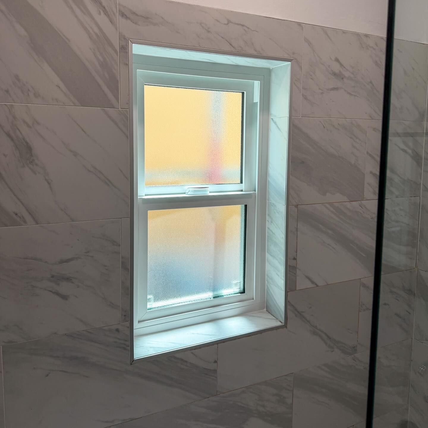 Small frosted window set in a gray tiled wall, with warm light glowing through the panes.