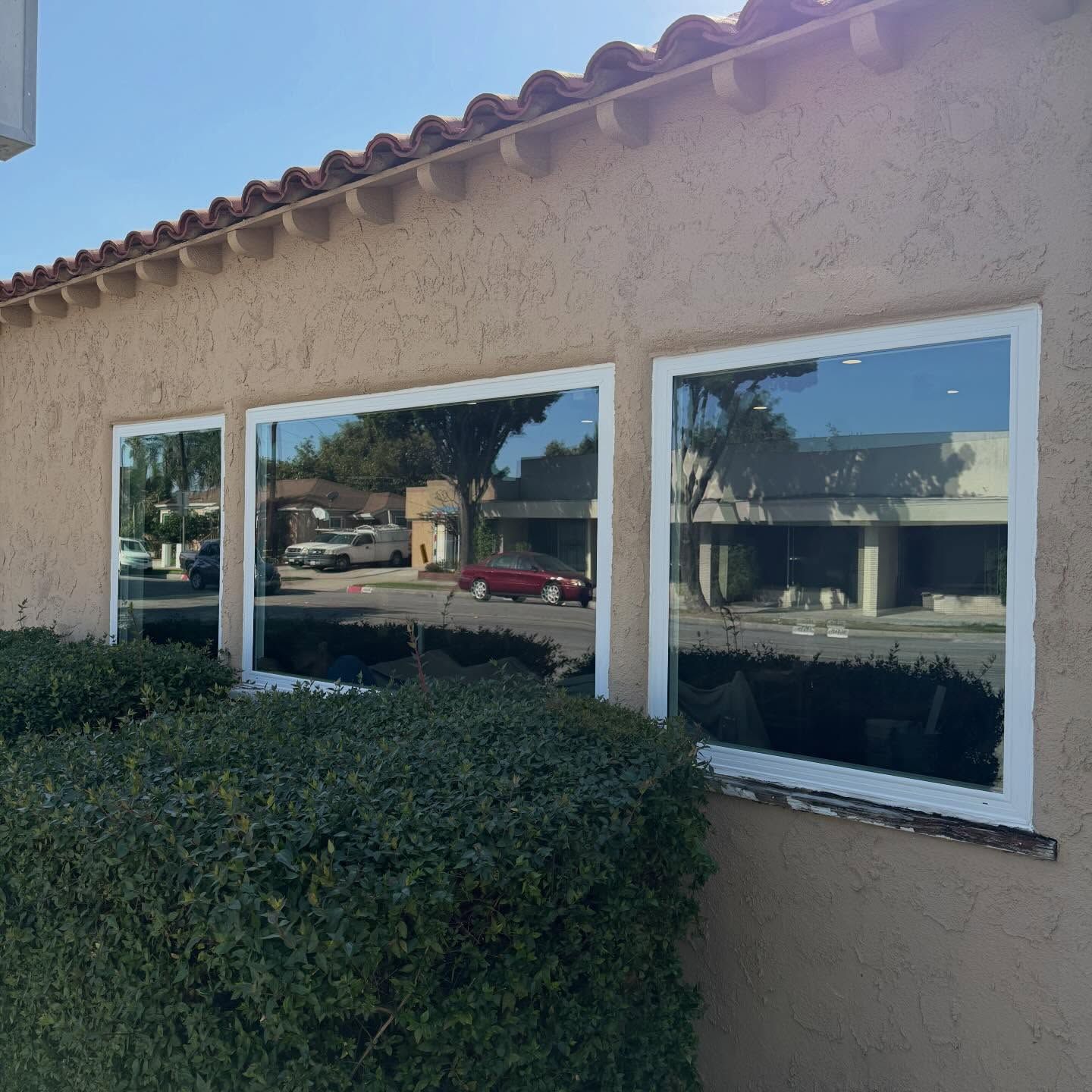 Tan stucco building with three windows and trimmed hedges in front, reflecting a parking lot and nearby buildings.