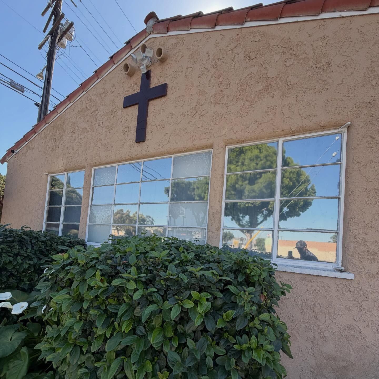 Beige building with a dark cross above windows, partly covered by bushes under a clear sky