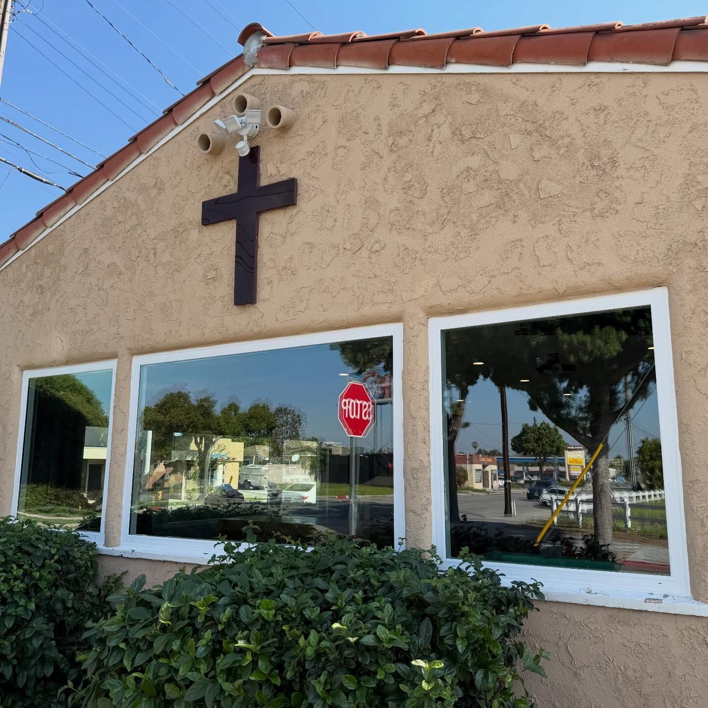 Small stucco building with a dark cross above windows, reflecting a street scene and a stop sign.