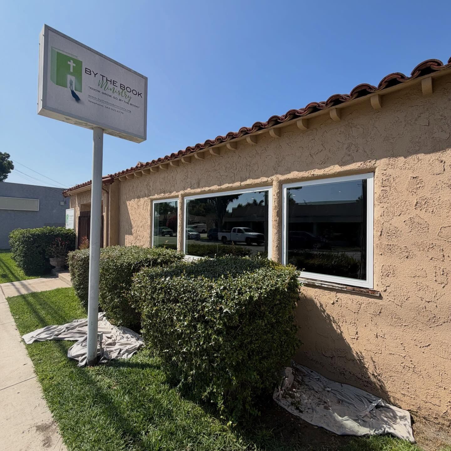 Sunlit stucco building with windows, hedge, and a roadside sign by the sidewalk