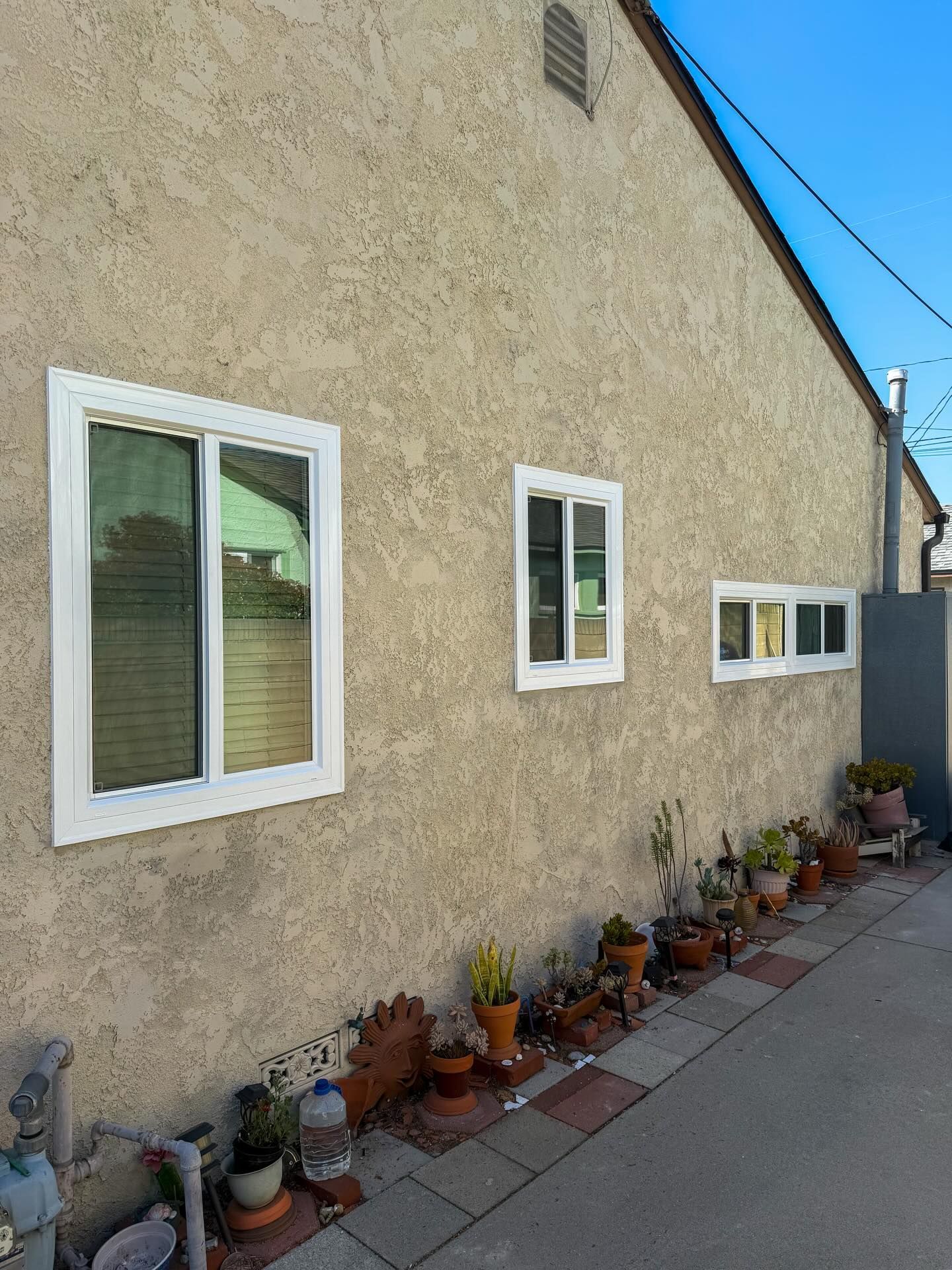 Stucco house wall with white windows and potted plants along a sidewalk under a blue sky
