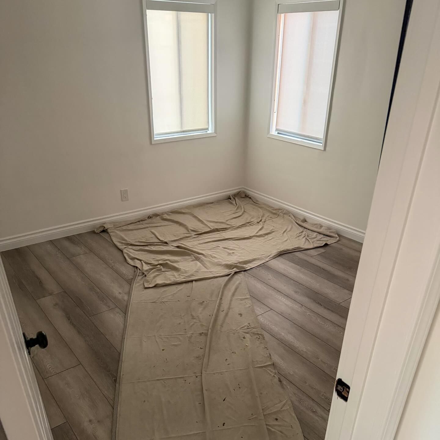 Empty small room with white walls, two windows, wood floor, and drop cloths laid on the floor