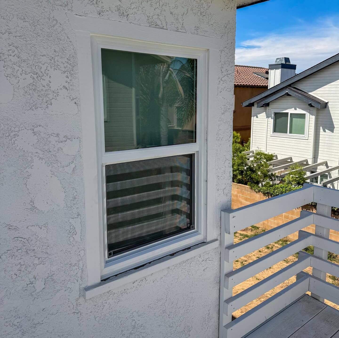 White house exterior with a window and balcony railing overlooking neighboring homes and blue sky