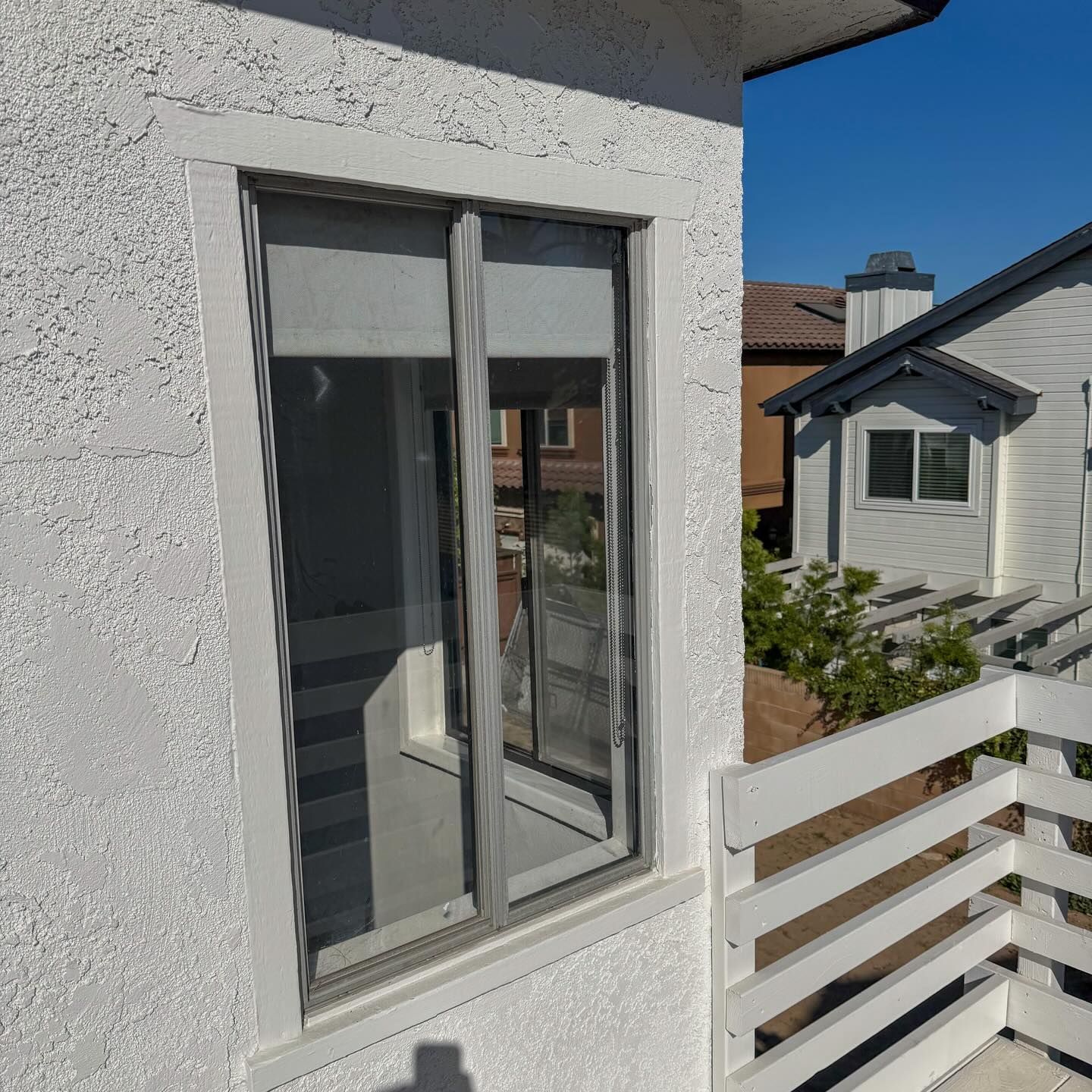 Sliding window on a white stucco wall beside a balcony railing, with neighboring houses in view.