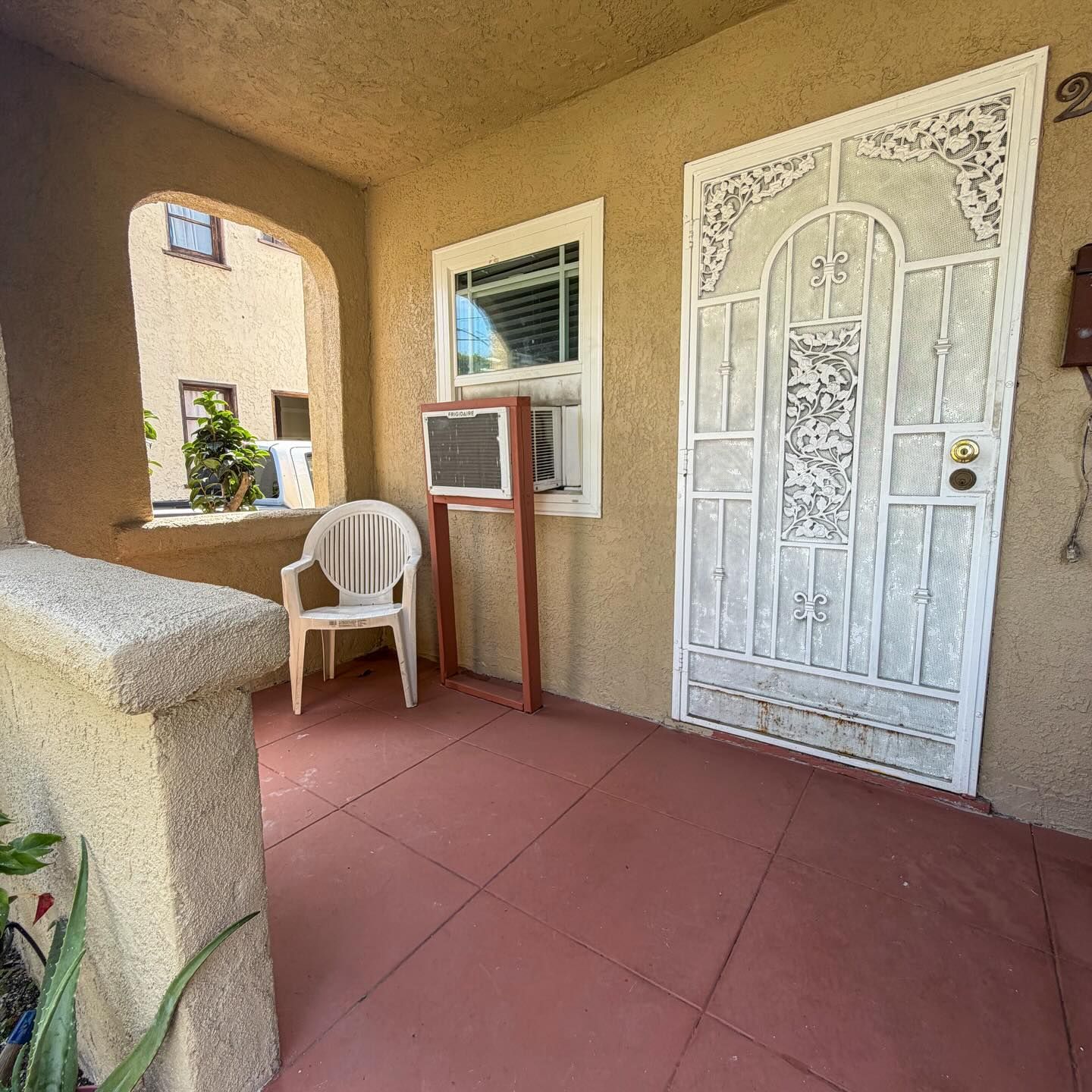 Front porch with white chair, window AC unit, and ornate white door on a red tiled floor