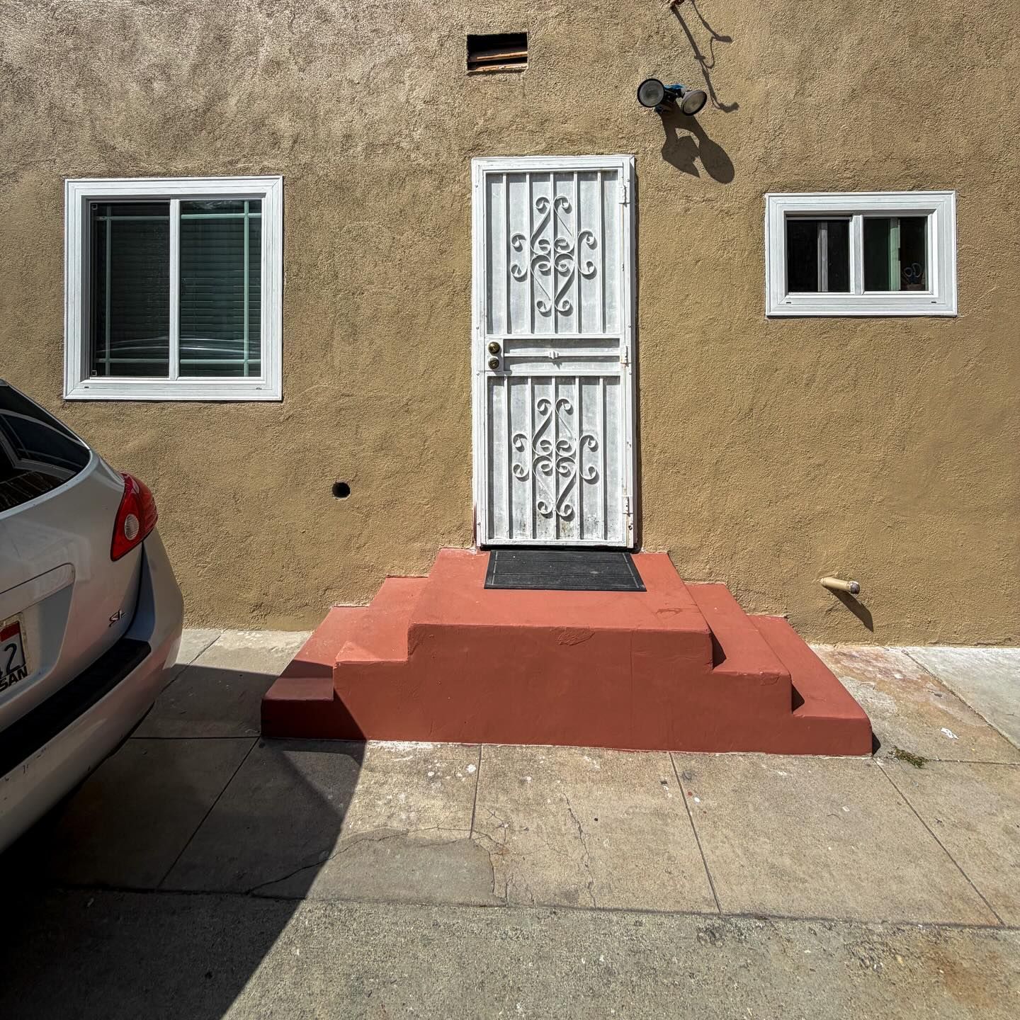 Front entrance of a tan house with a white security door, small red steps, and two windows; a car is parked at left.