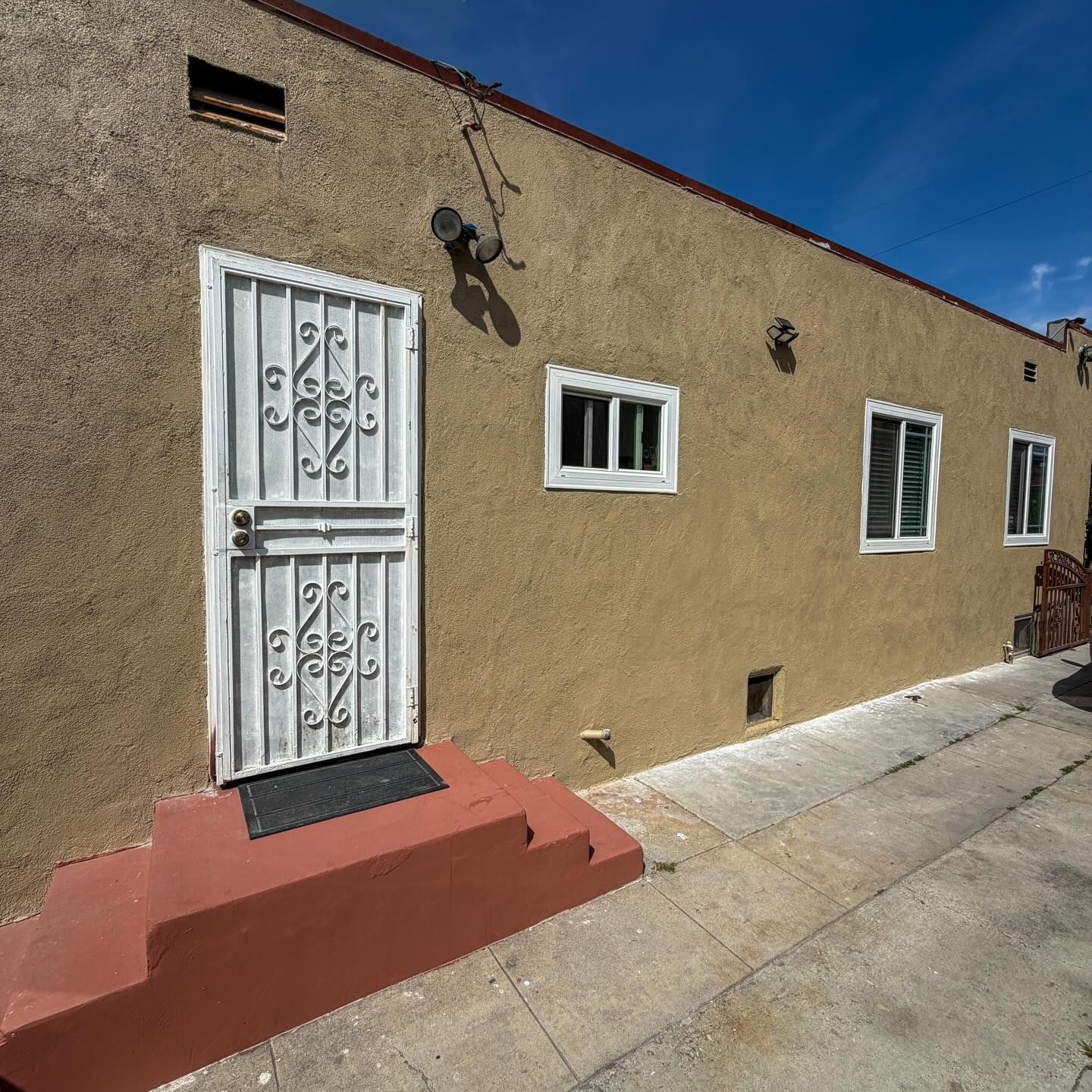 Sunlit stucco house wall with white barred door, small windows, and a concrete sidewalk