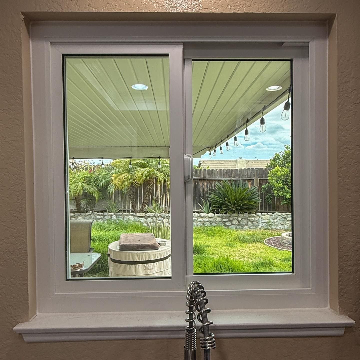 Kitchen window looking onto a backyard patio with a round table and palm trees.