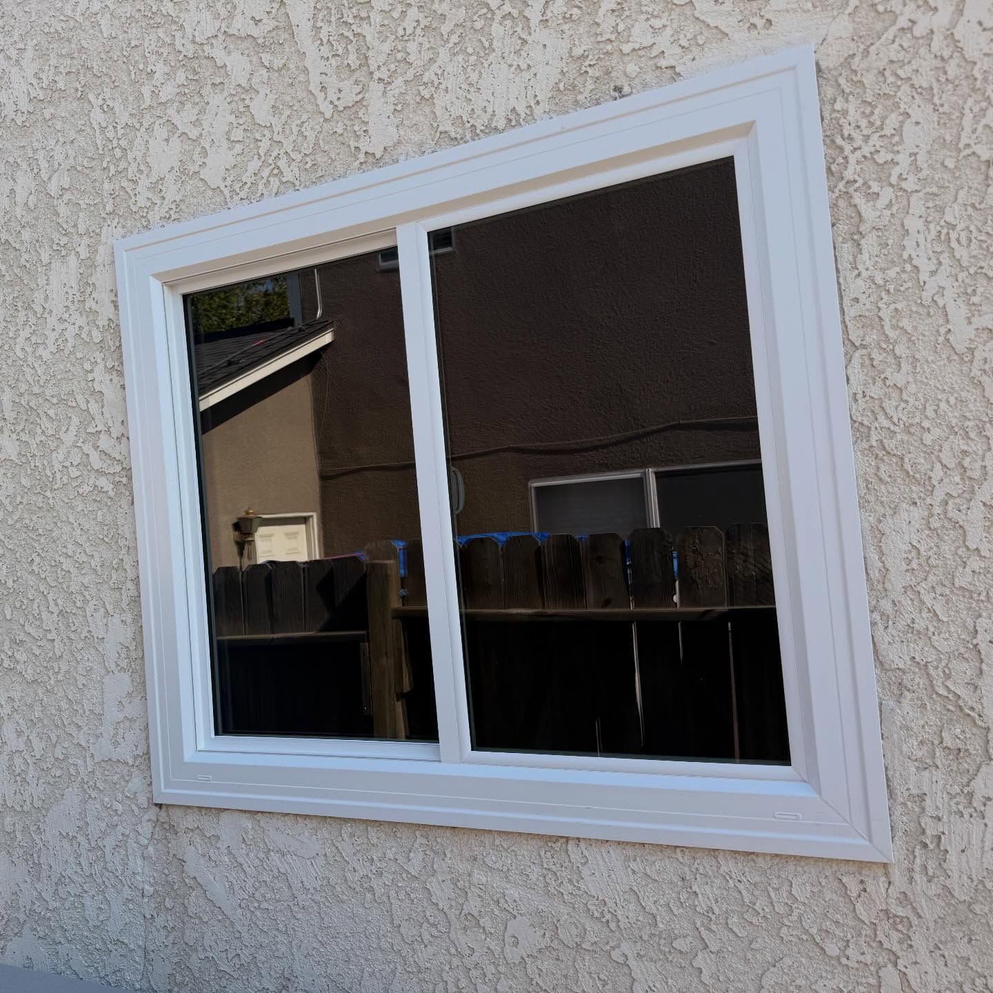 White-framed sliding window in a beige stucco wall, reflecting a patio and house across the way
