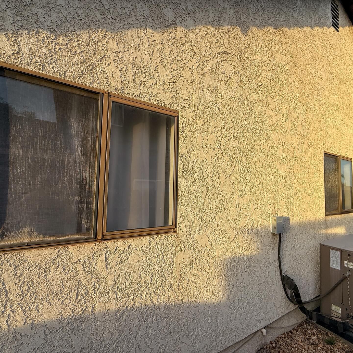 Sunlit stucco house wall with two windows, an exterior light, and an air conditioning unit.