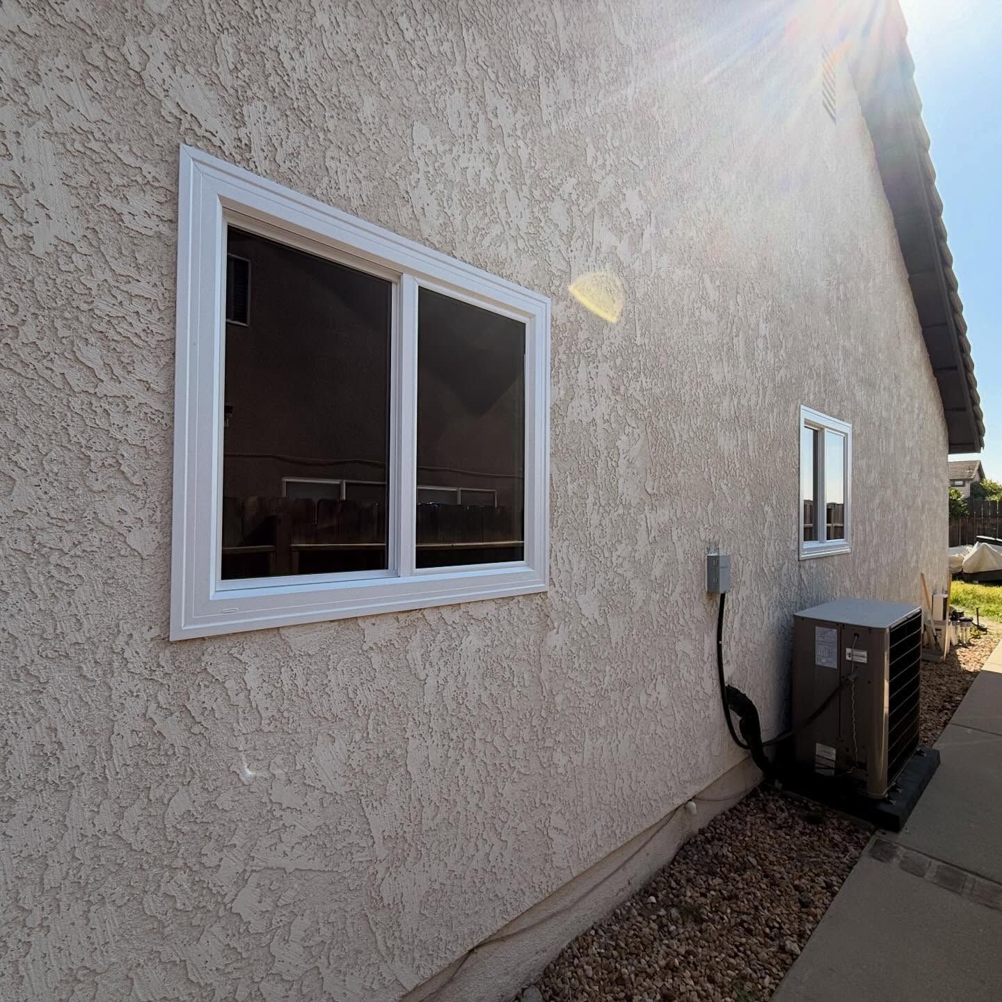 Stucco house exterior with two windows, gravel border, and an air-conditioning unit along the wall.