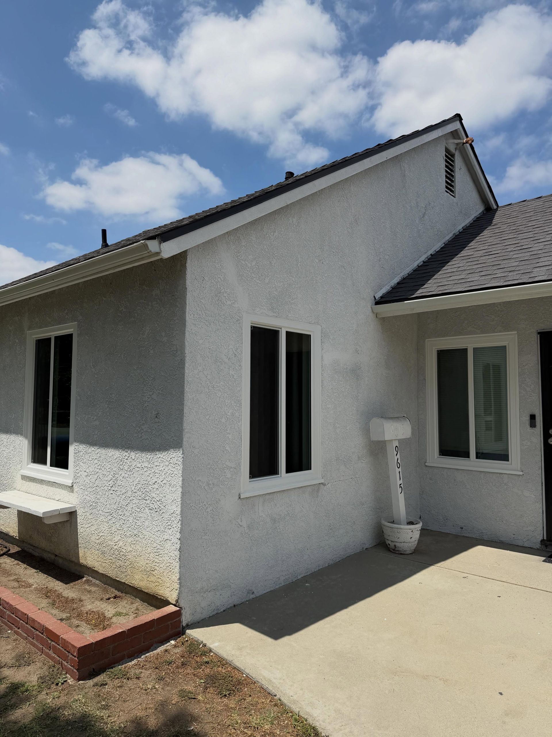 White stucco house exterior with black-trimmed windows, gray roof, and a sunny concrete patio.