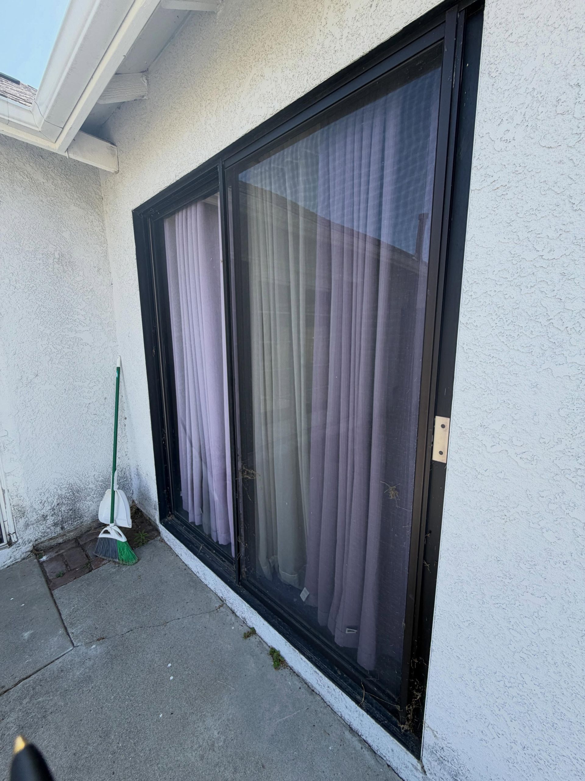 Sliding glass patio door with sheer curtains on a stucco exterior wall, beside a concrete walkway.