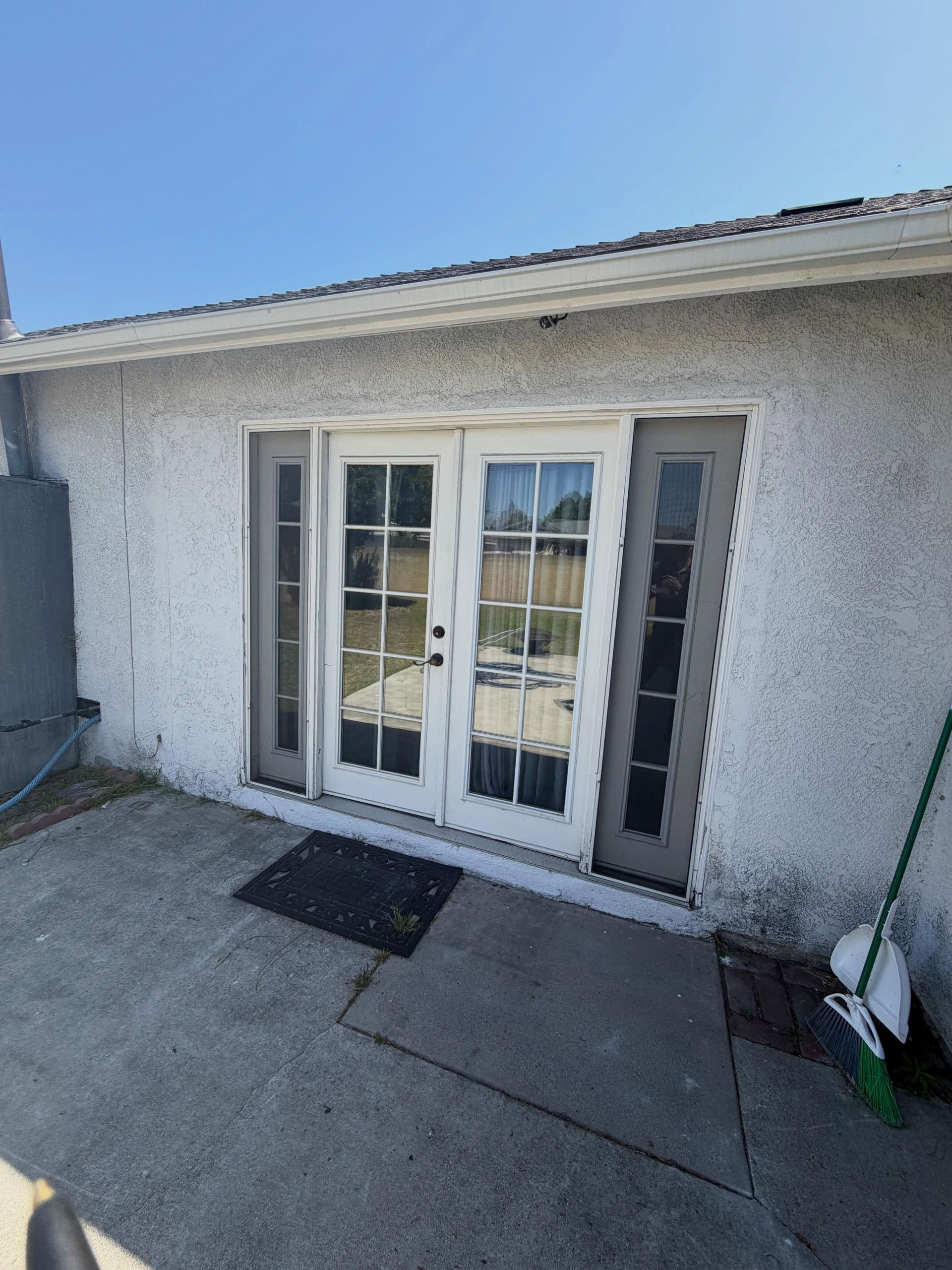 White stucco house exterior with double glass doors and a narrow side window, viewed from a concrete patio