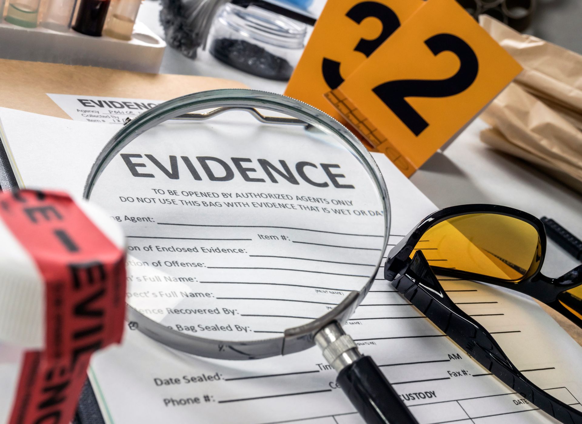 Evidence file under magnifying glass with sunglasses, pen, and yellow “2” marker on desk