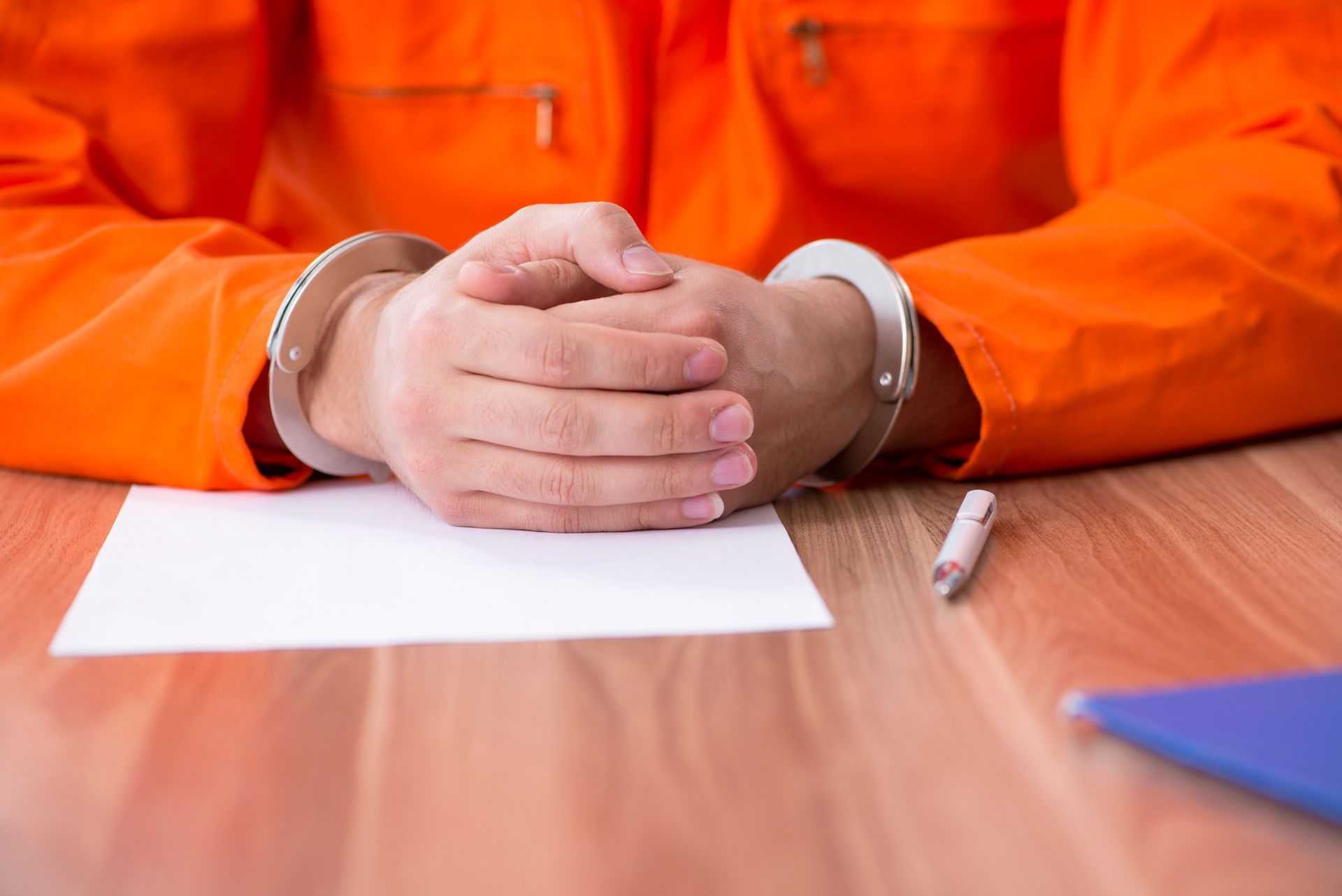 Person in orange jumpsuit with handcuffed hands on a desk, next to paper and pen.