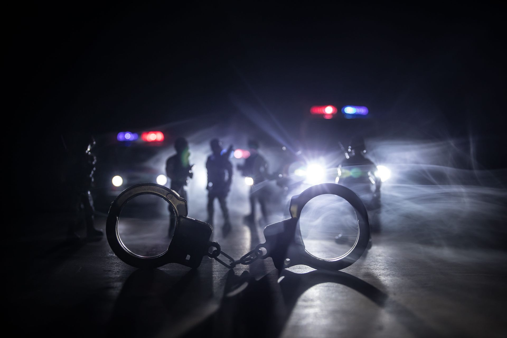 Handcuffs in foreground with police cars and officers in flashing lights at night