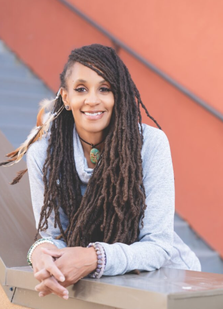 Smiling person leaning on a railing, with long dreadlocks and a gray sweater against an orange wall.