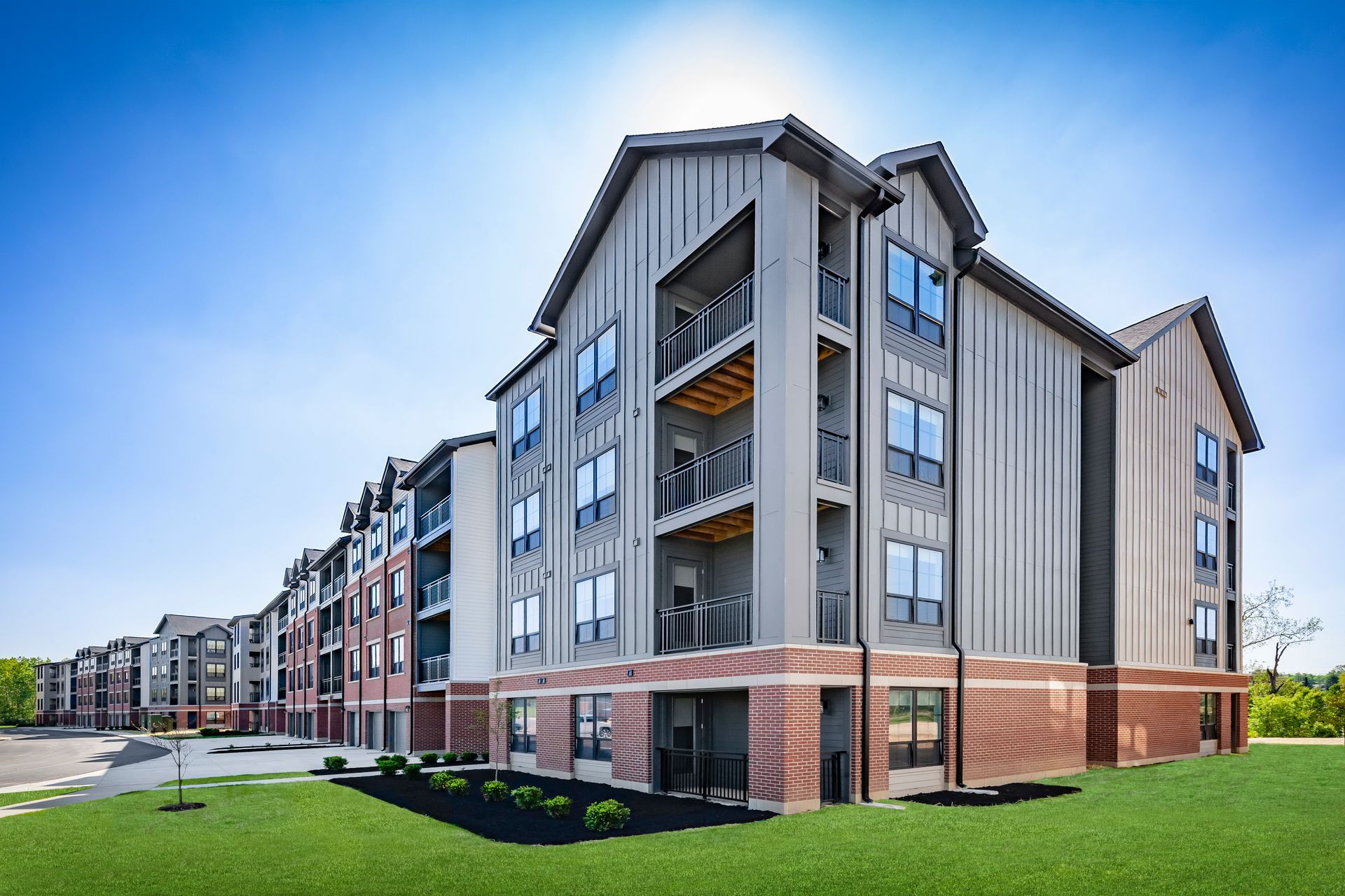 Modern apartment building with vertical gray siding, brick base, and balconies. Sunny day with clear blue sky at River Place Flats in Fishers, IN.