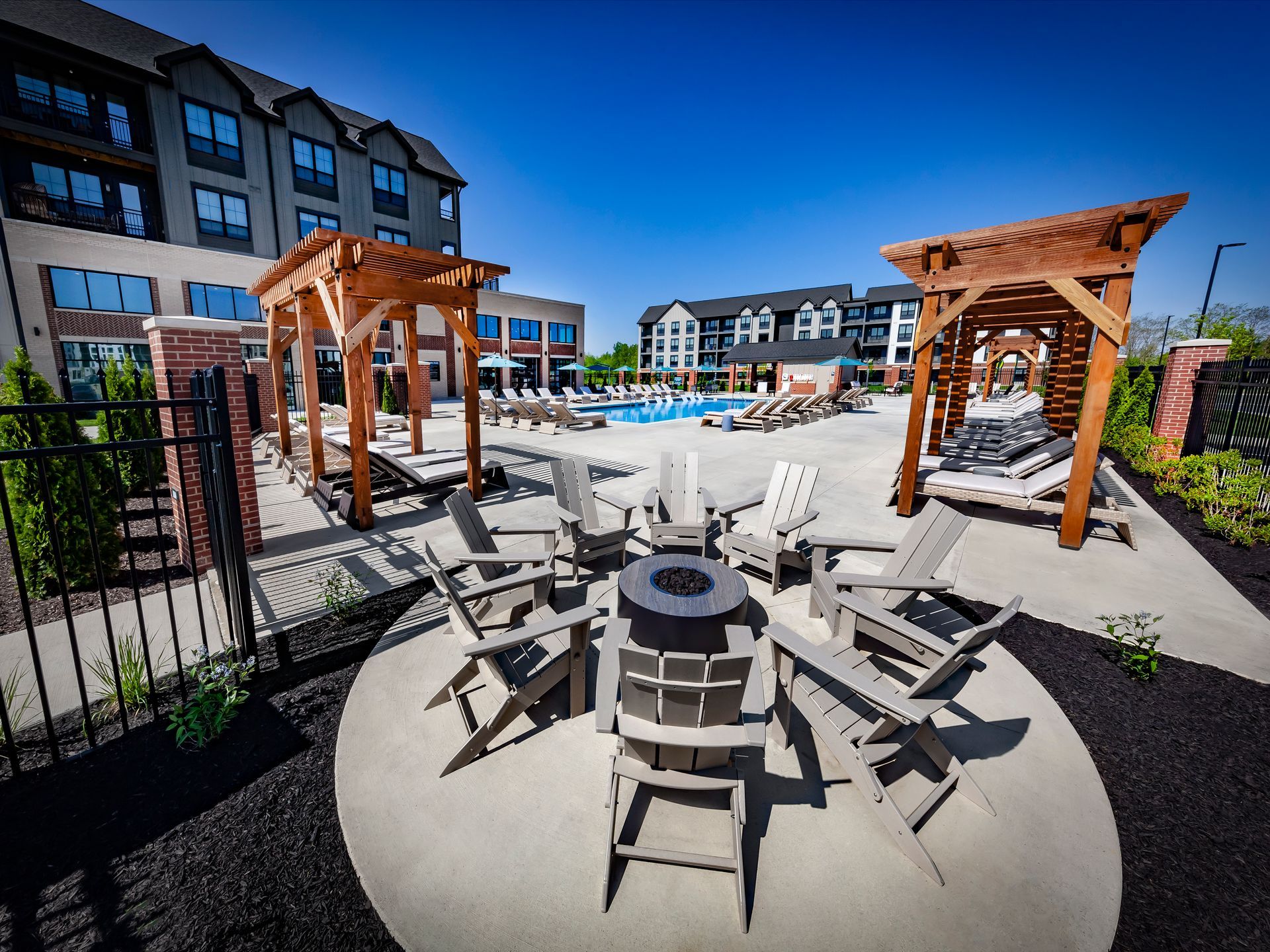 Outdoor lounge area with fire pit, seating, pergolas, and pool; apartment buildings in background at River Place Flats in Fishers, IN.