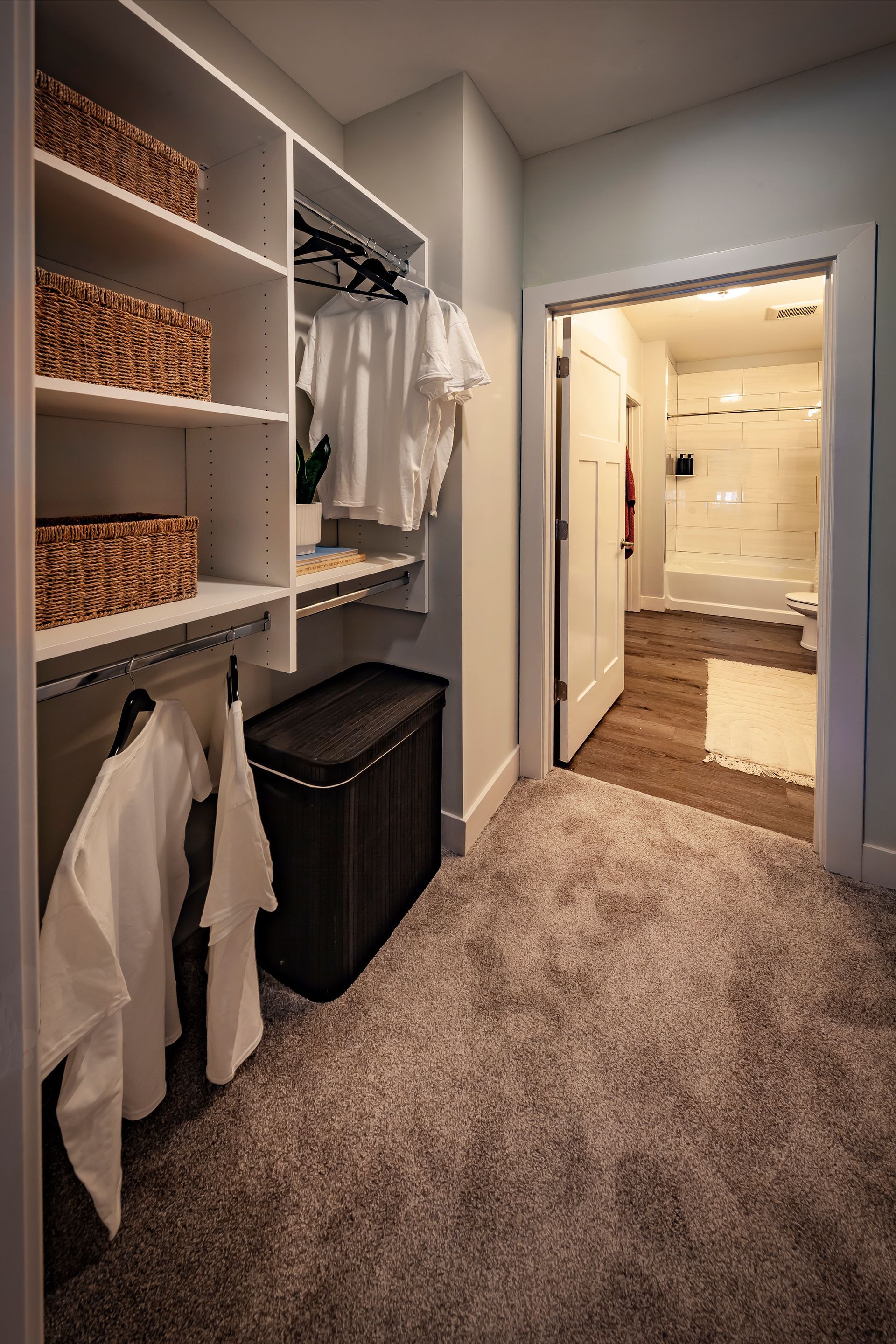 Walk-in closet with built-in shelves, hampers, and a doorway to a bathroom with a bathtub; carpeted floor at River Place Flats in Fishers, IN.