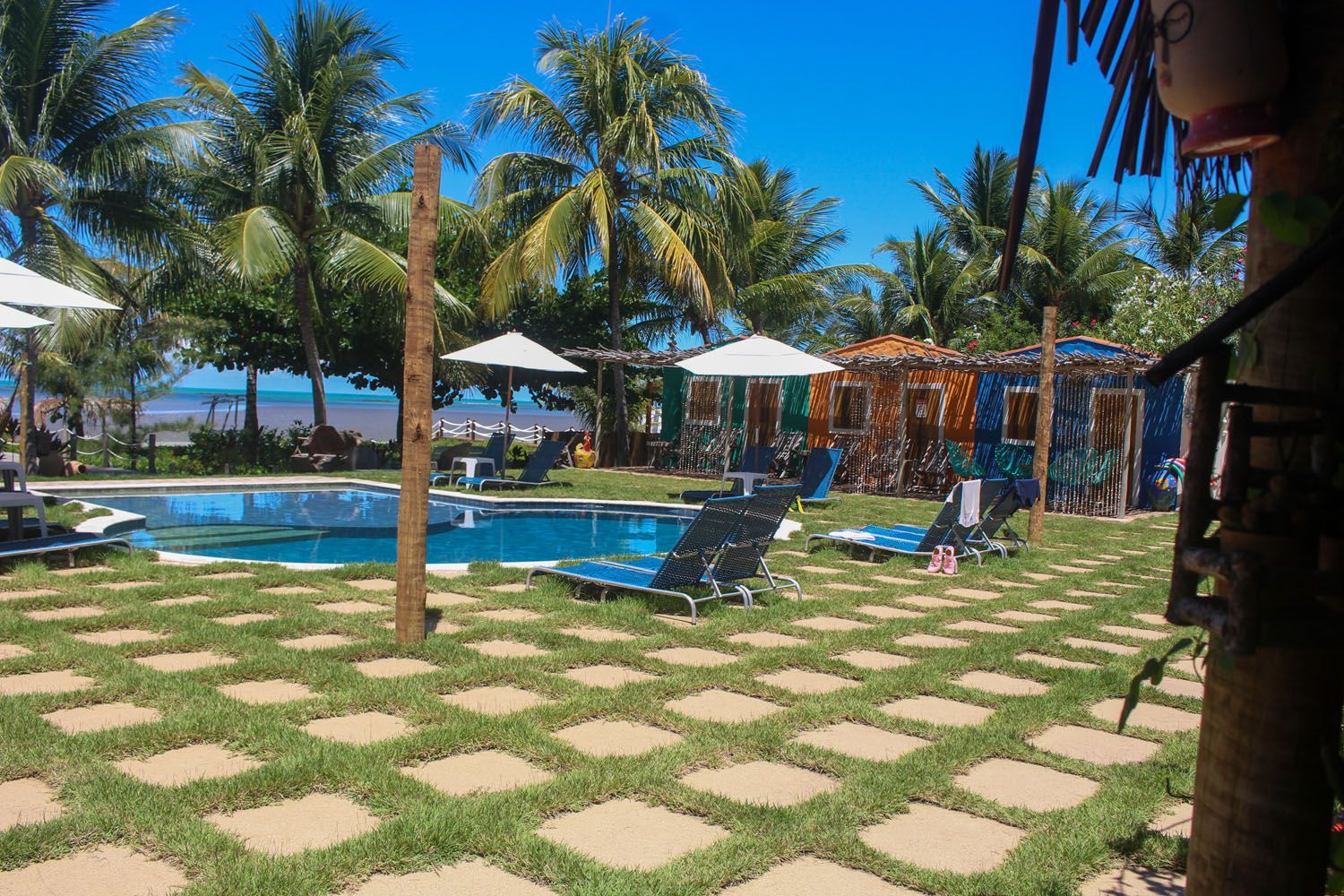Cenário à beira da piscina com espreguiçadeiras, praças de grama e areia, palmeiras e cabanas coloridas.