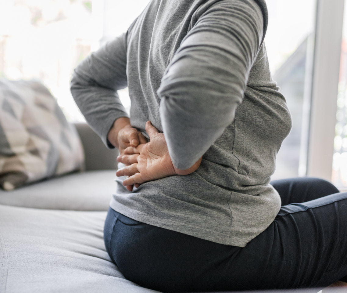 A woman is sitting on a couch holding her back in pain.