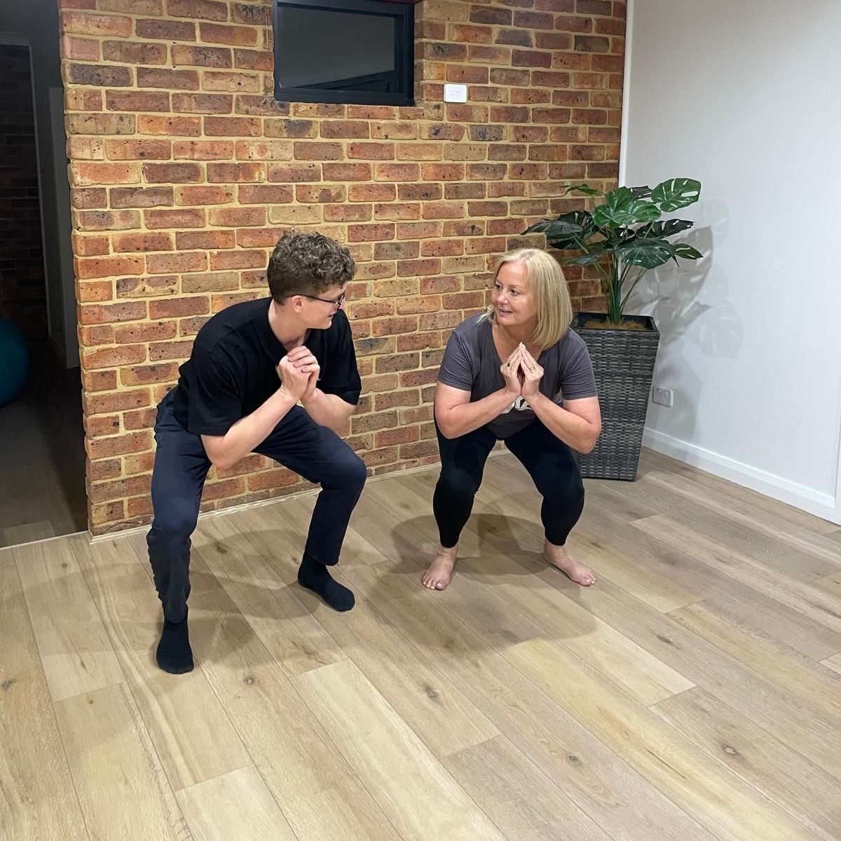 A physiotherapist in Sydney and a client are squatting on a wooden floor in a room.