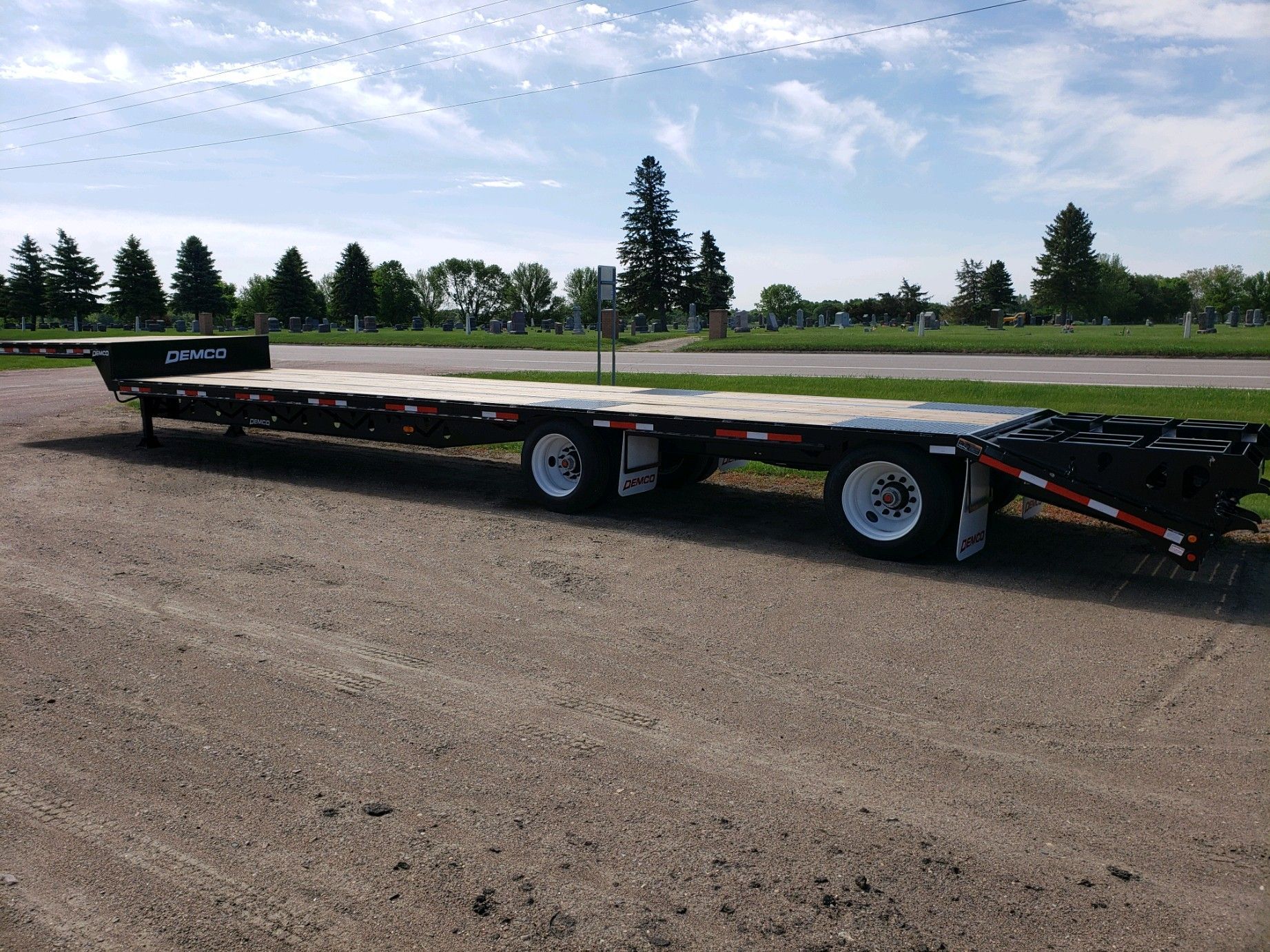 Flatbed trailer parked on gravel, with a wooden deck and white wheels, in front of trees.