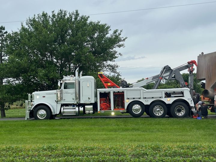 White tow truck with extended boom lifting a structure, parked on grass near a road and tree.