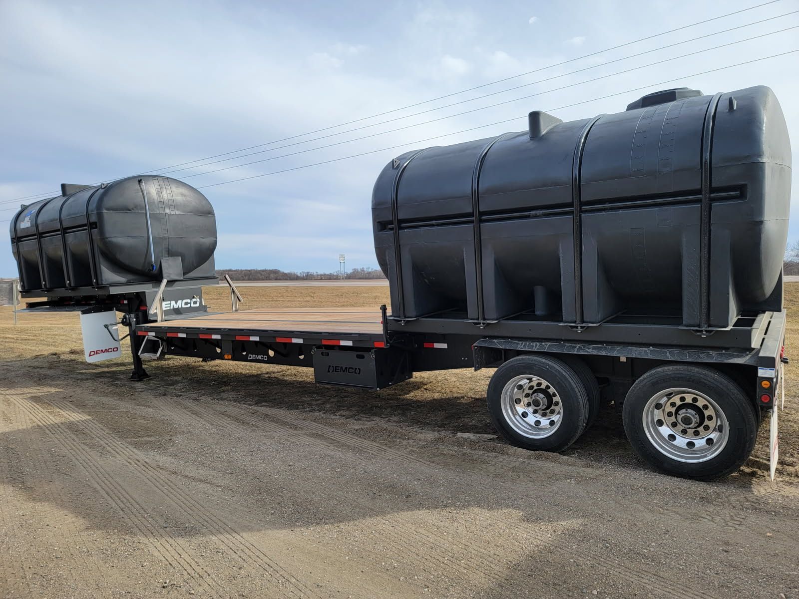 Black water tanks on a flatbed trailer on a dirt road, with a cloudy sky in the background.