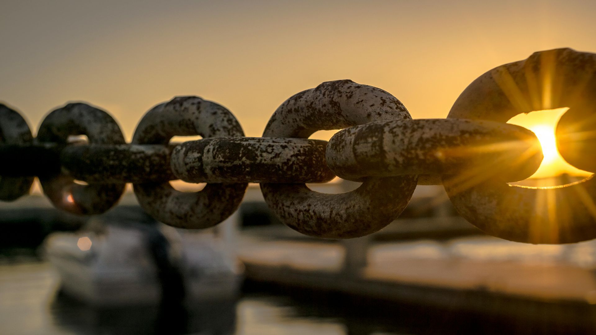 Rusty chain links with the sun shining through one; blurred dock in background.