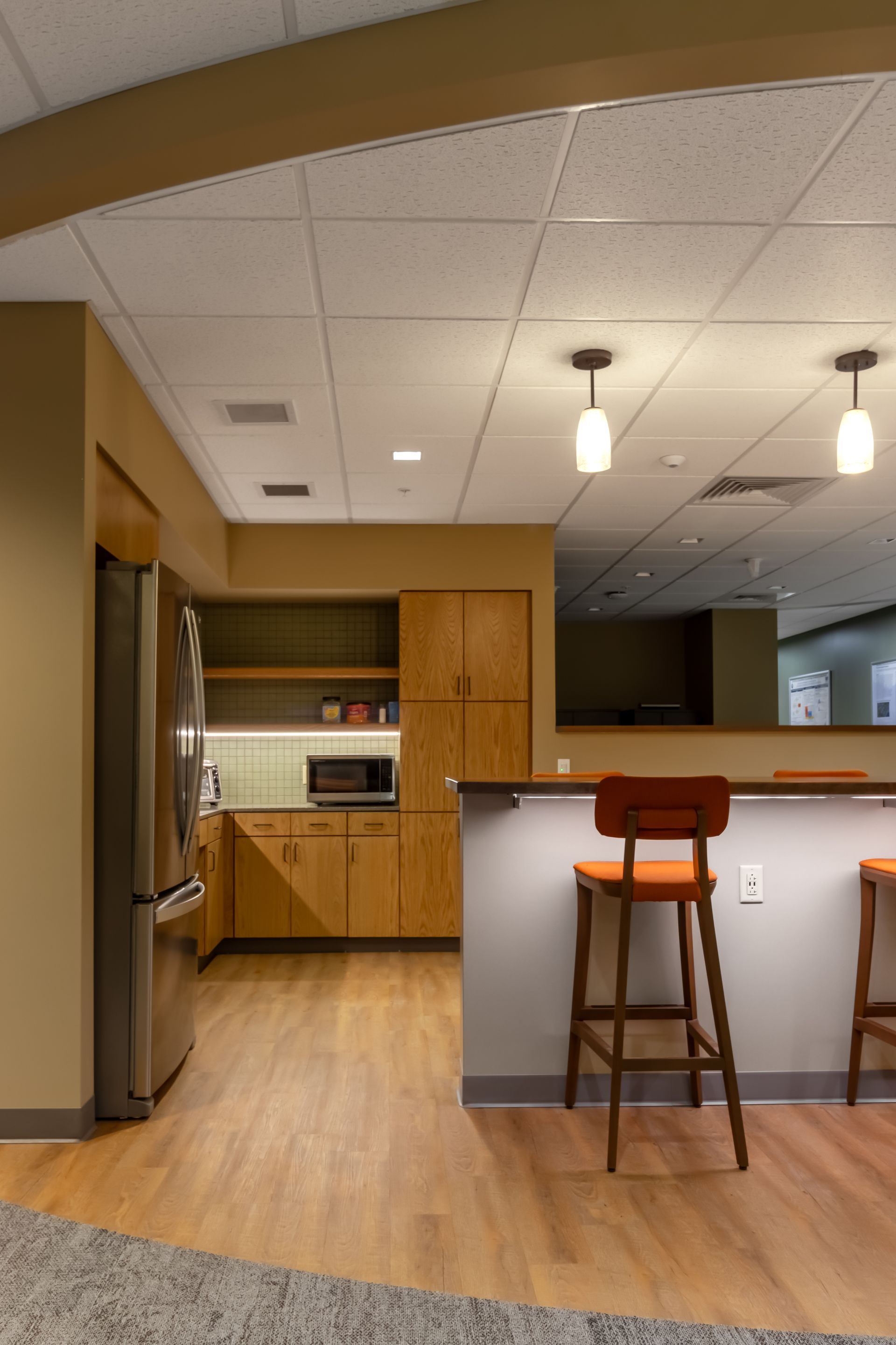 Penn State EESI Suite's kitchen with stainless fridge, wooden cabinets, white island, and two bar stools under pendant lights.