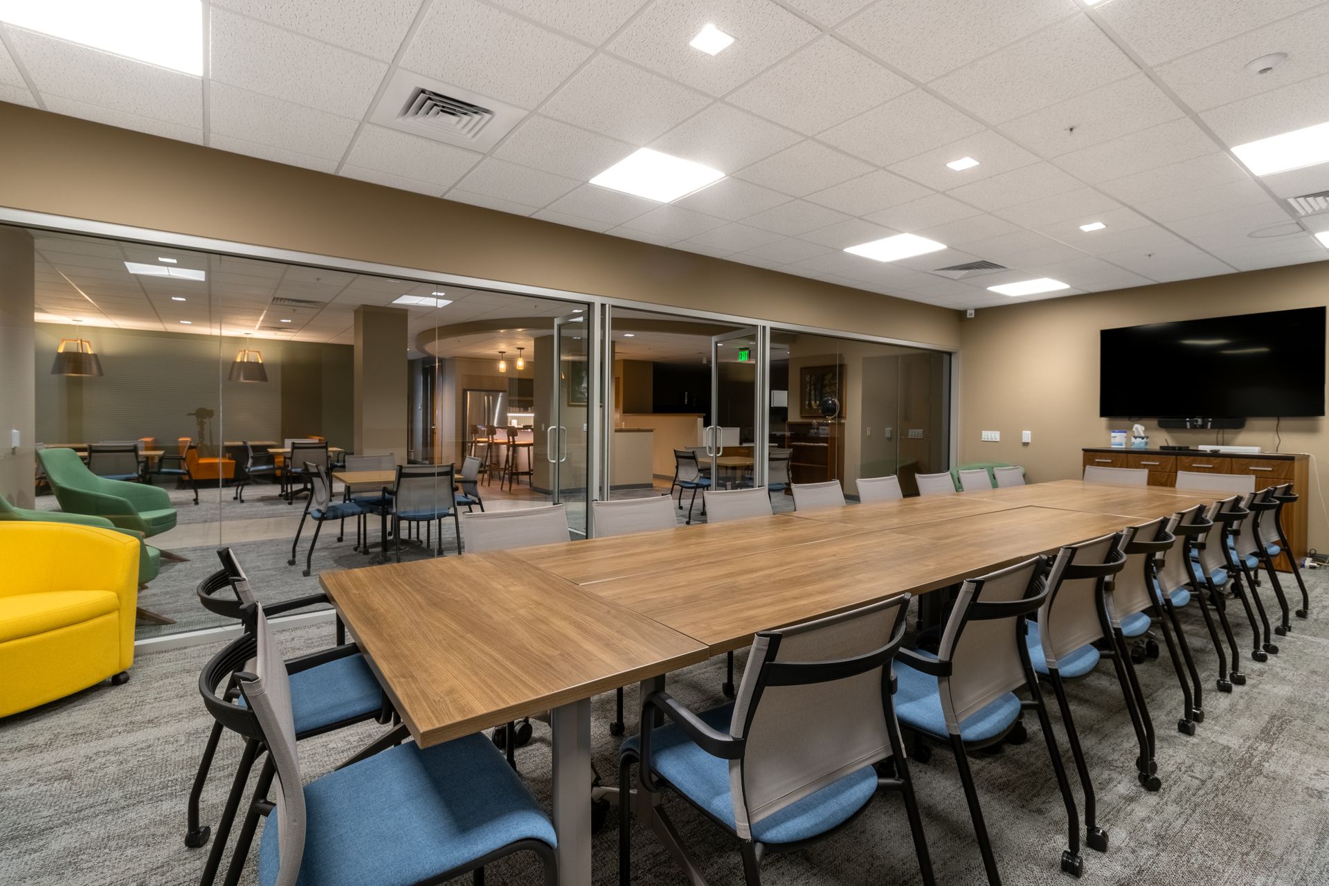 Modern conference room with long wooden table, blue chairs, yellow sofa, and wall-mounted TV
