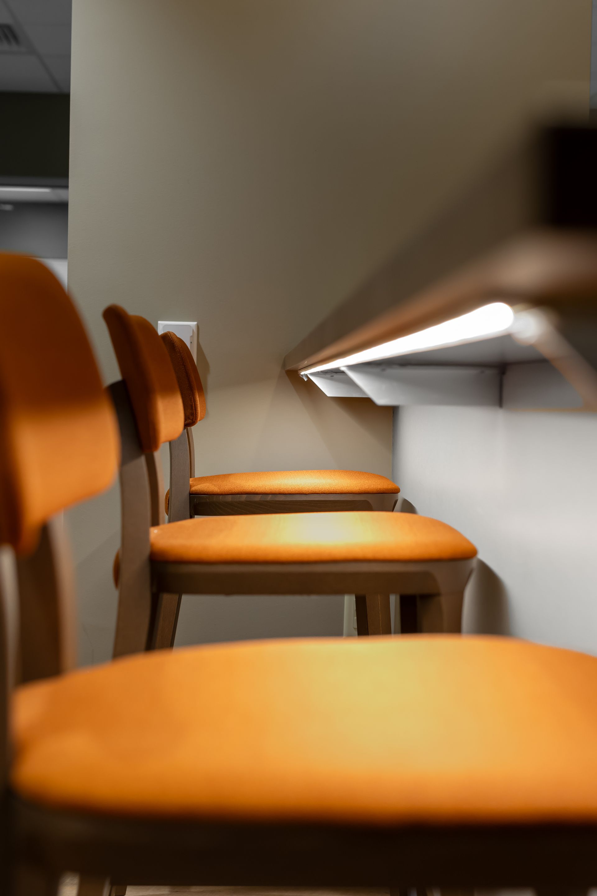 Row of orange chairs beside a wall-mounted light in a narrow hallway or waiting area