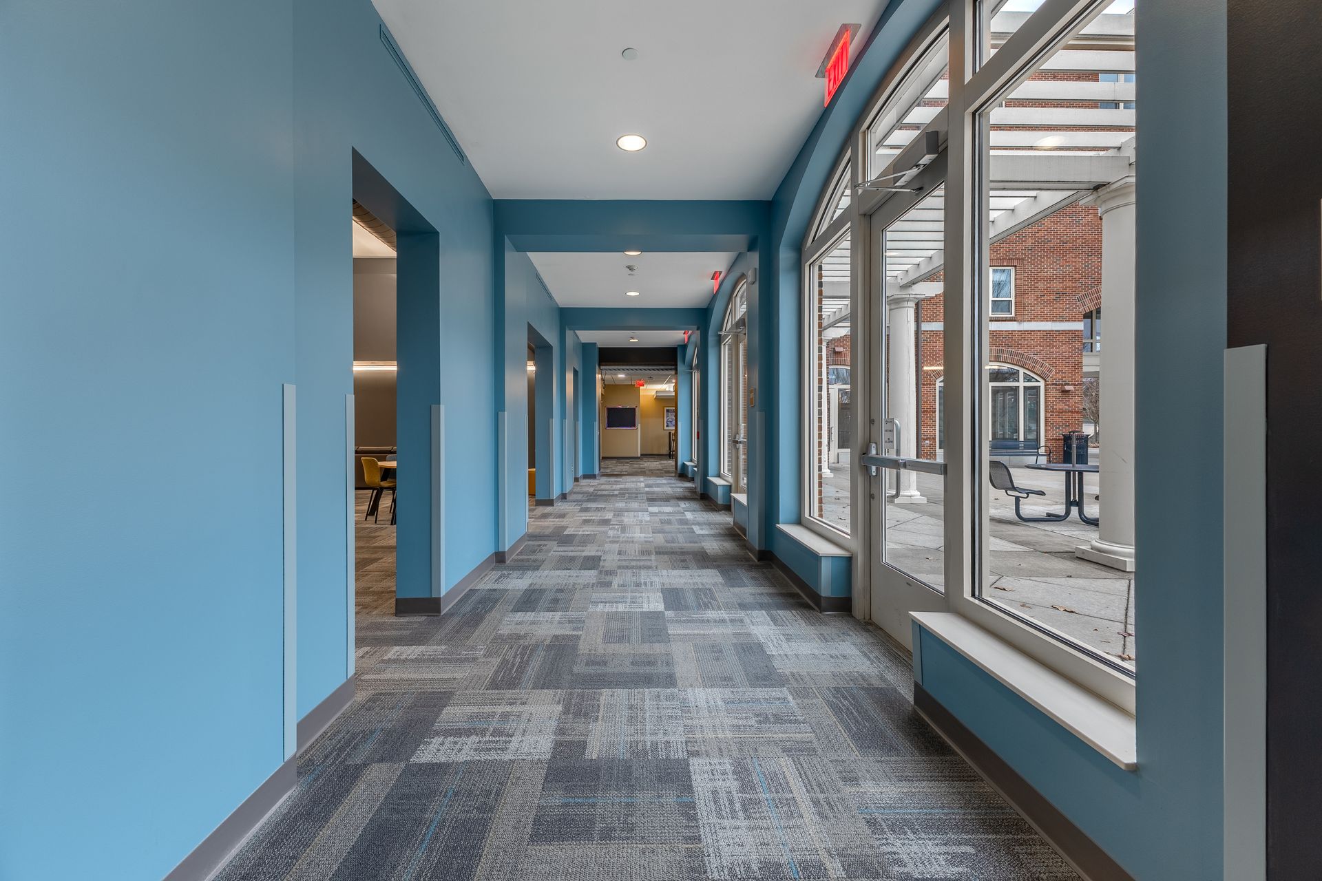 Eastview Terrace Kitchen's modern hallway with blue walls, carpeted floor, and large windows overlooking a brick building.